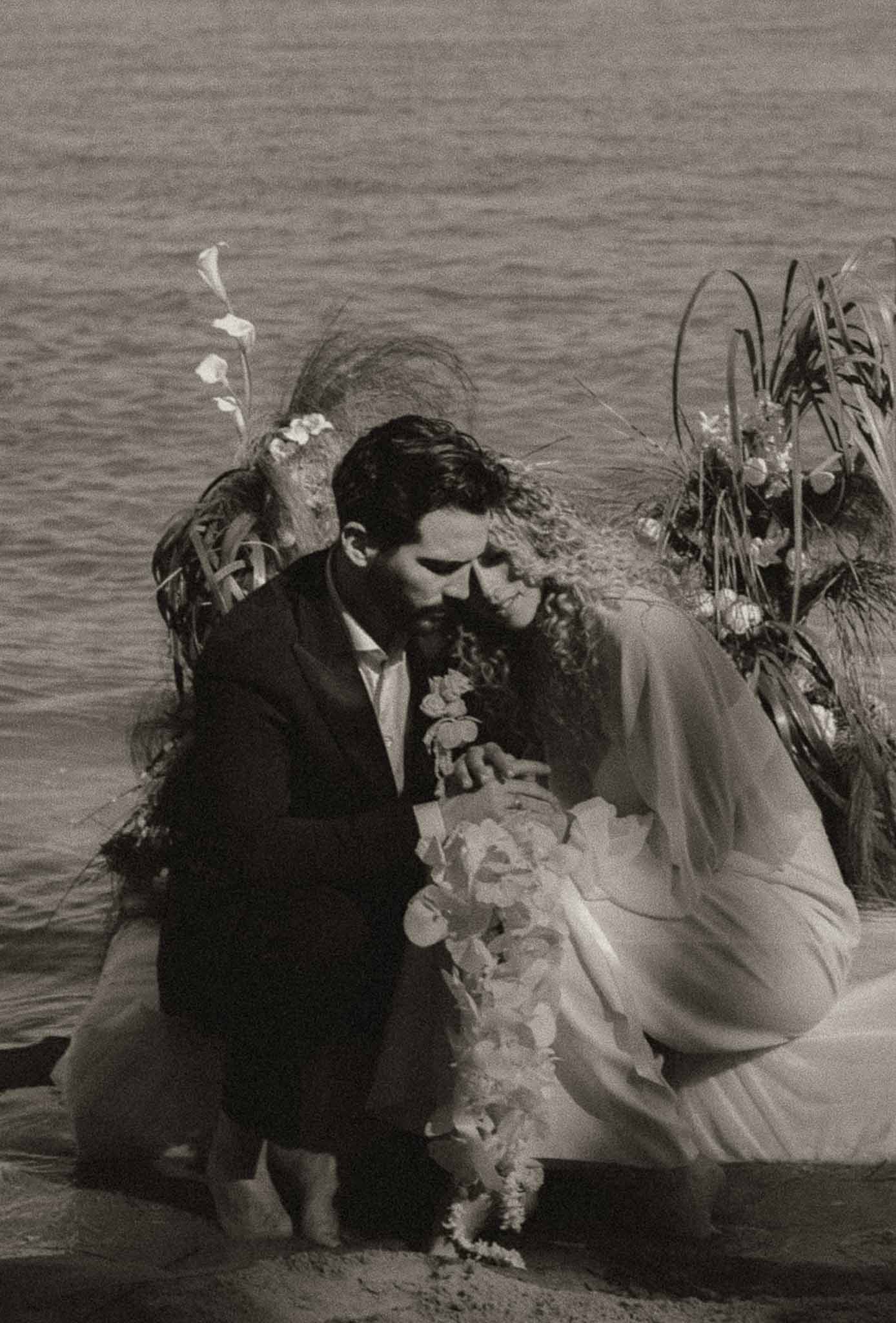 Couple crouches at waterside with cascading bouquet beside calla lily installation in B&W film grain