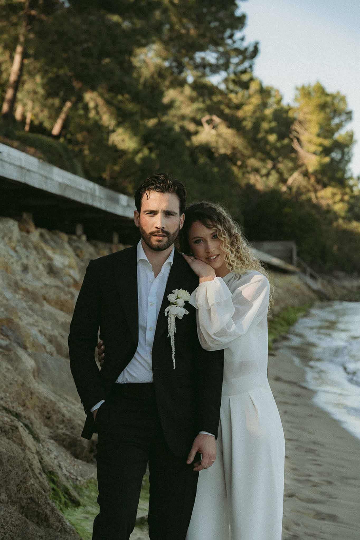 Couple on sandy beach at golden hour, bride in sheer bishop sleeve ivory dress leaning against groom in black suit