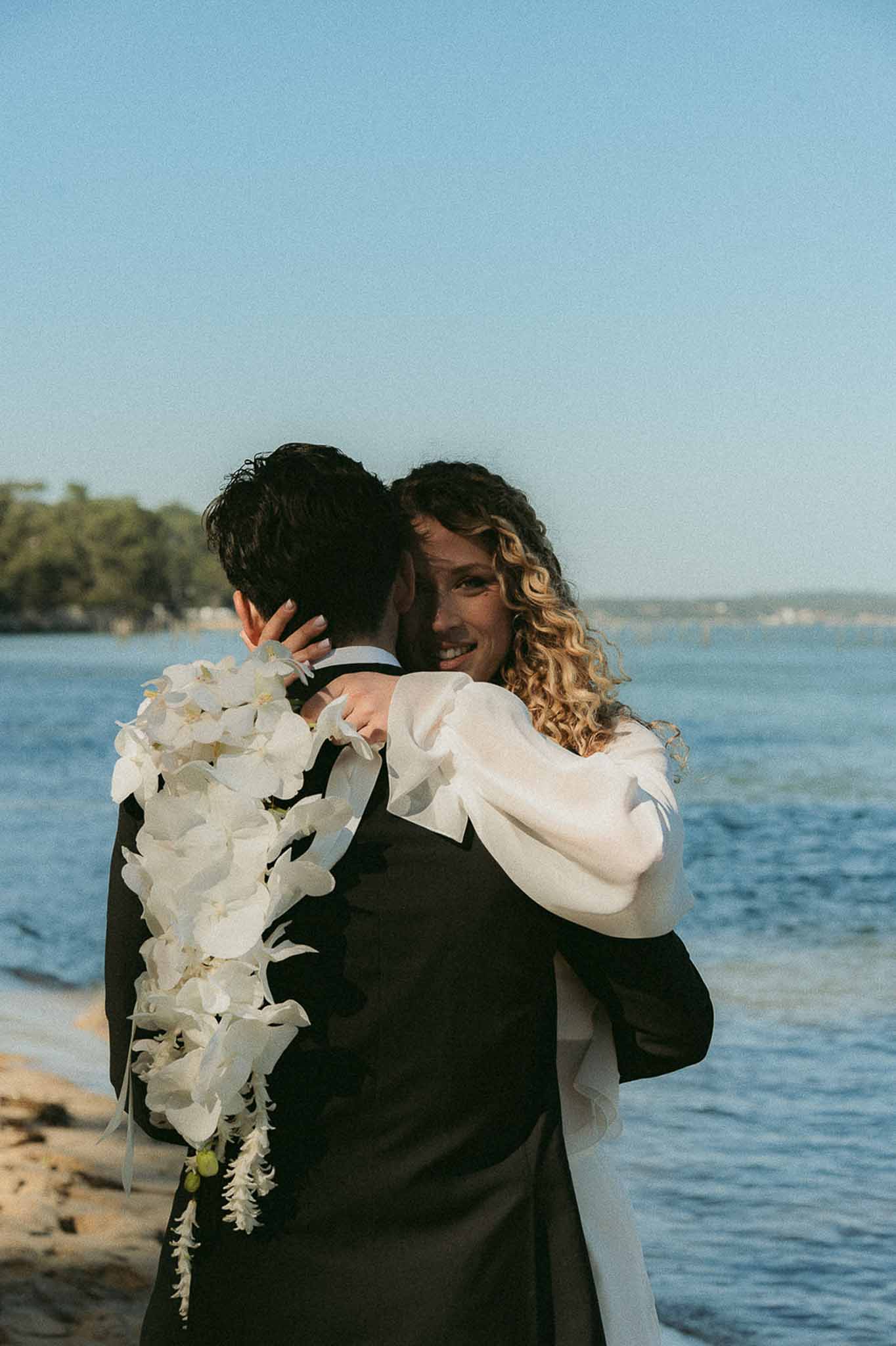 Bride and groom embracing on a sandy beach, bride holding a cascading white phalaenopsis orchid bouquet