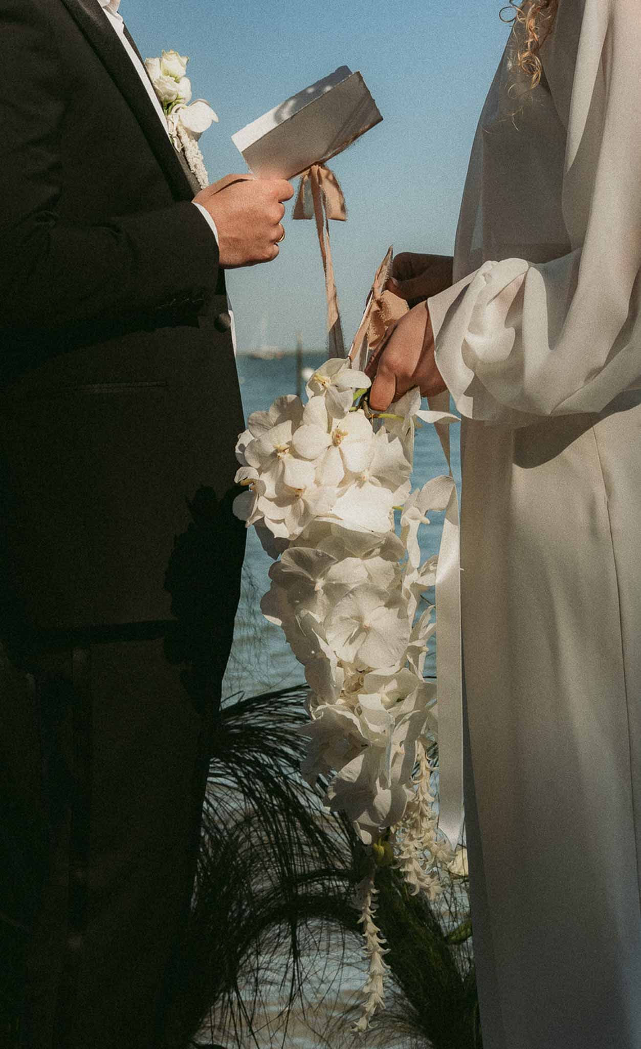Close-up of couple at waterside ceremony showing white orchid cascade bouquet and ceremony booklet