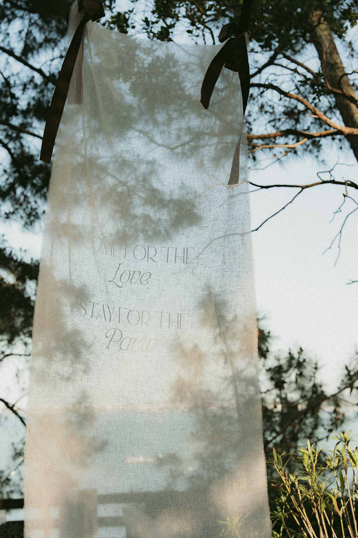 Sheer linen banner reading Come for the Love Stay for the Party hanging from tree branches outdoors