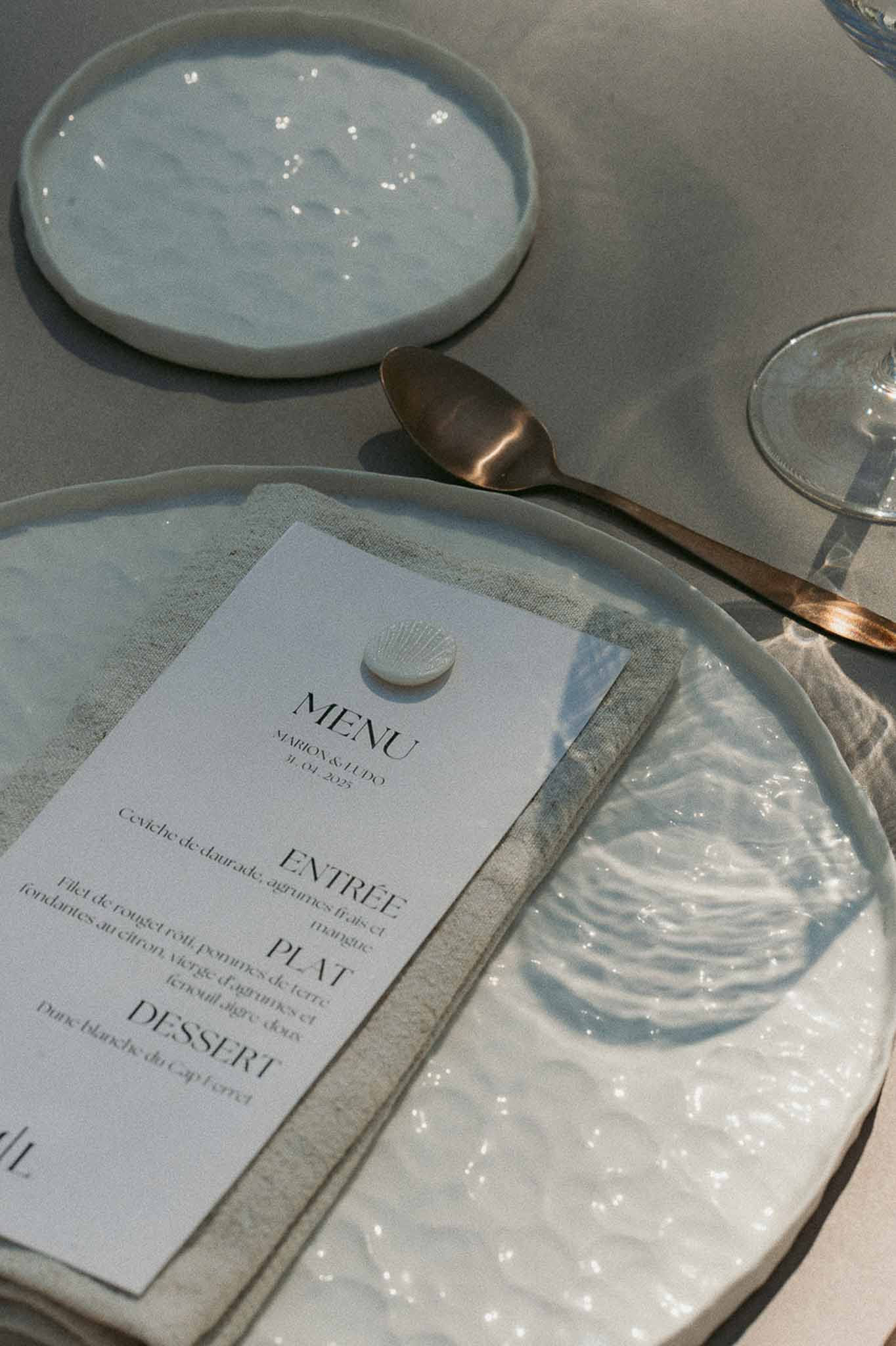 Coastal-inspired place setting with wavy glass charger plate, French menu card, and shell place card holder on grey linen
