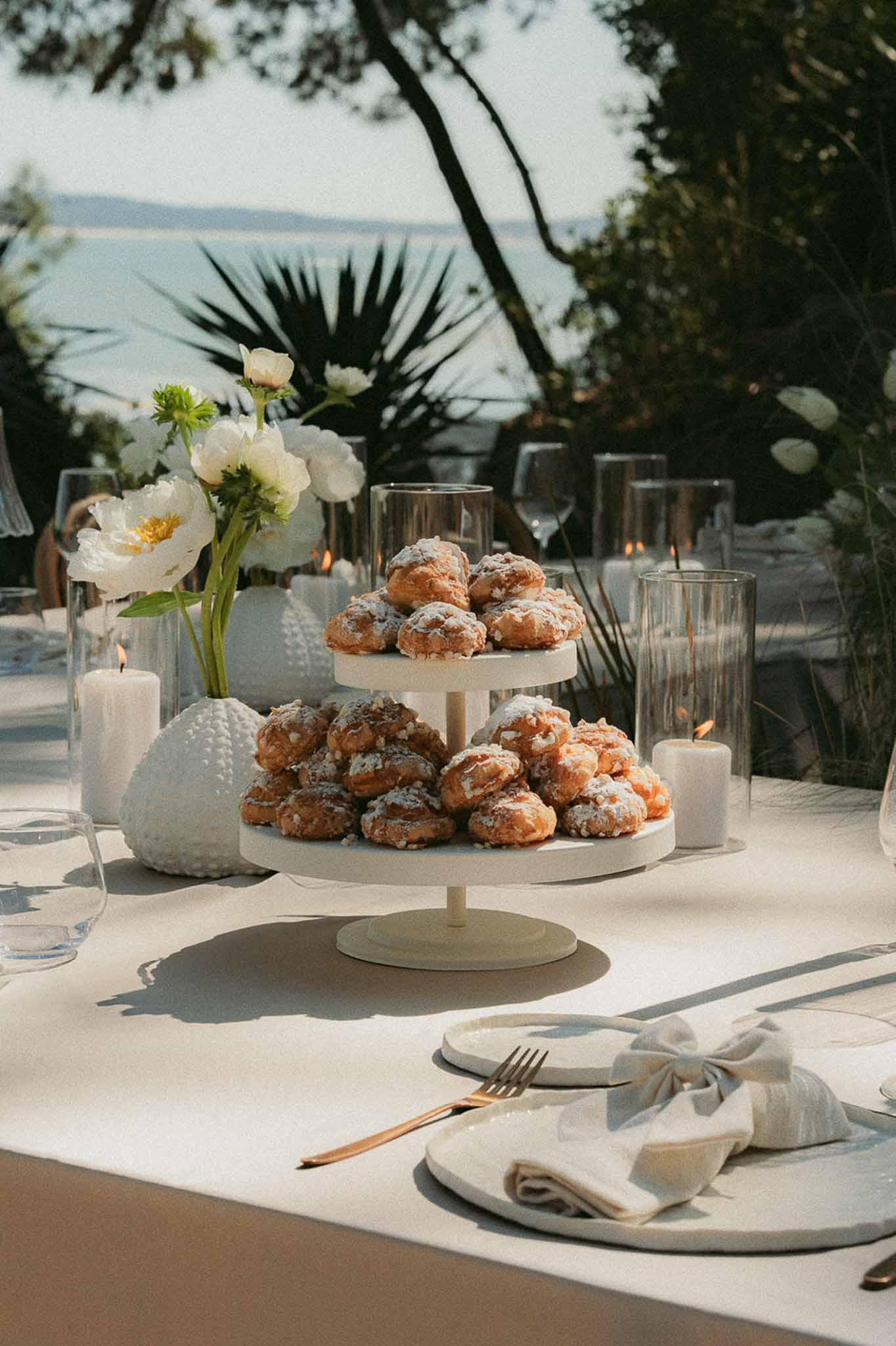 Choux pastry tower with white peony centerpiece, gold cutlery, and hurricane candles on coastal table