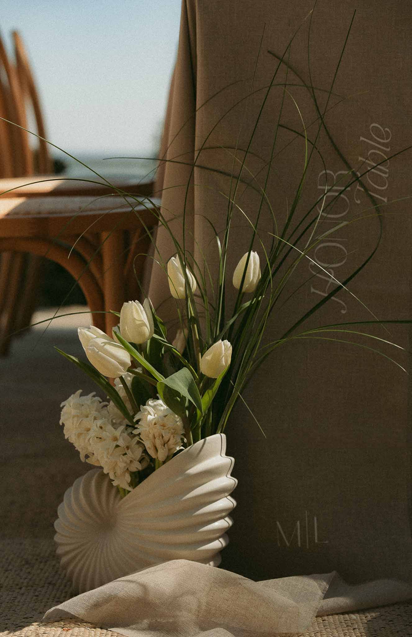 Shell-shaped ceramic vase with ivory tulips and hyacinths beside monogrammed chair cover at reception