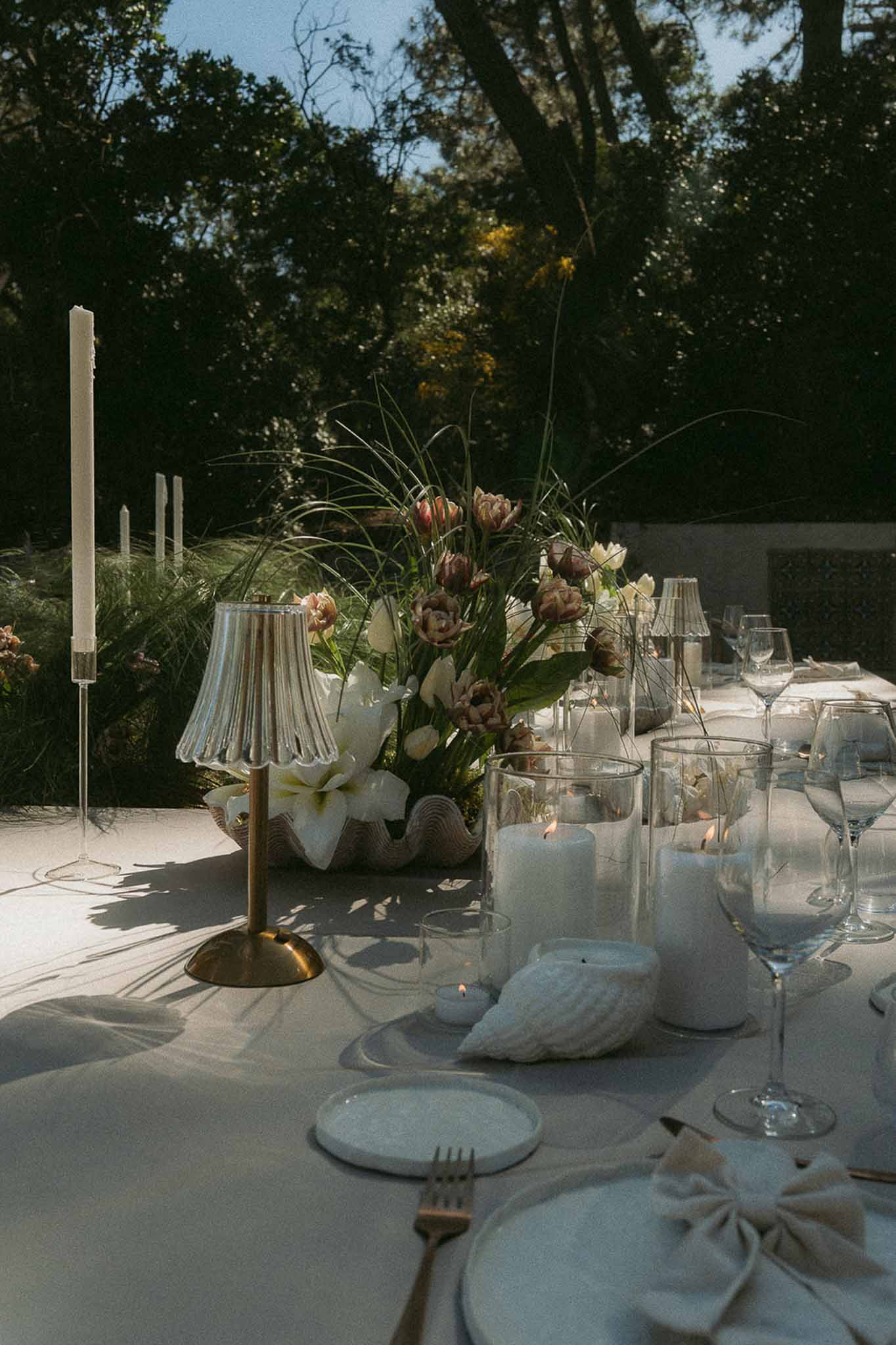 Coastal-styled place setting with shell vessel of mauve tulips, brass lamp, and ivory tapers in sunlight