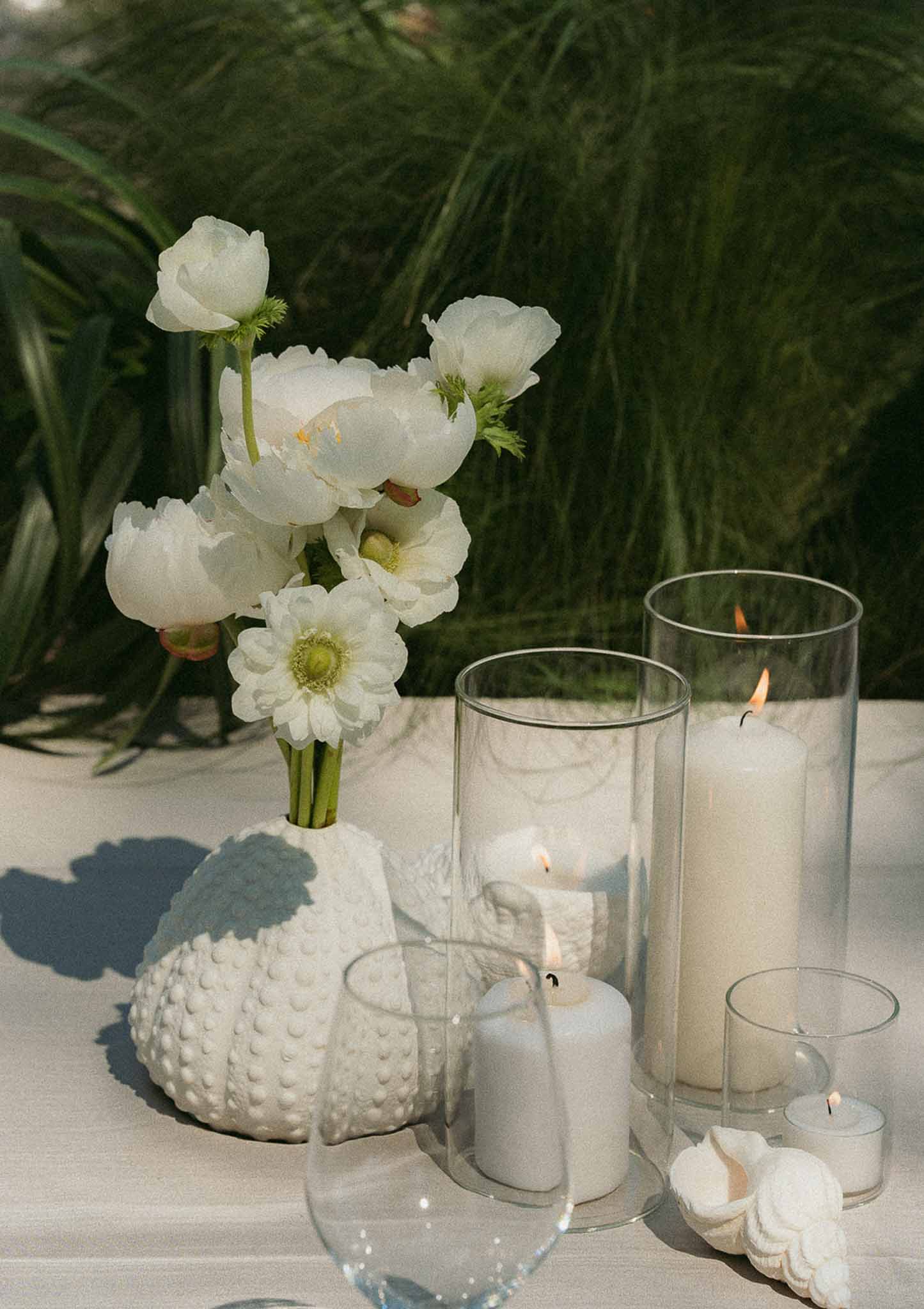 Sea-urchin vase with white peonies and ranunculus beside hurricane candles and seashells on white linen