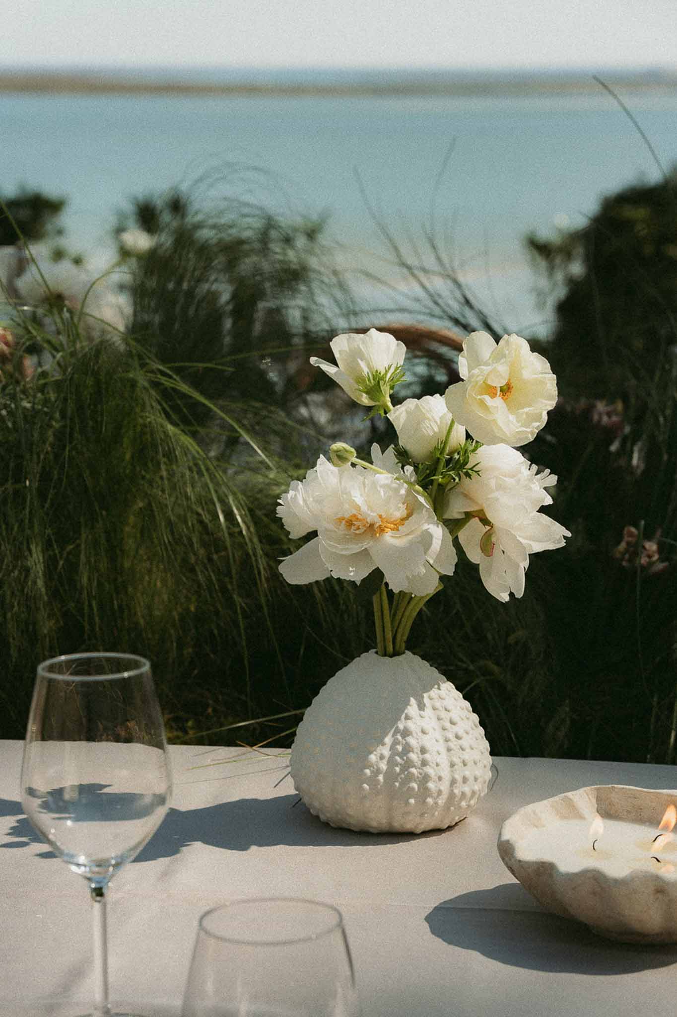 Coastal table setting with cream peonies in sea urchin vase, shell candle dish, and waterfront backdrop