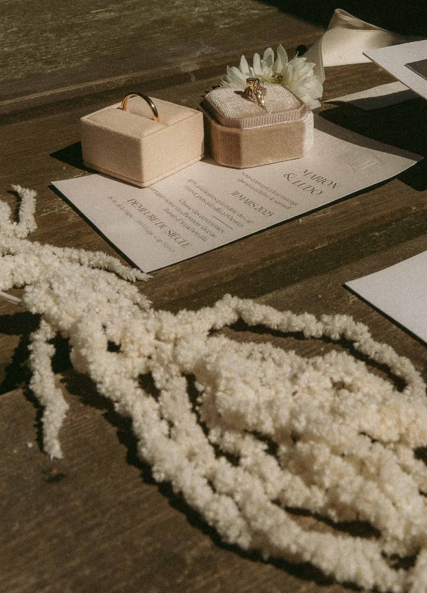 Flat lay of wedding invitation, gold rings in velvet boxes, and dried flowers on dark wood surface