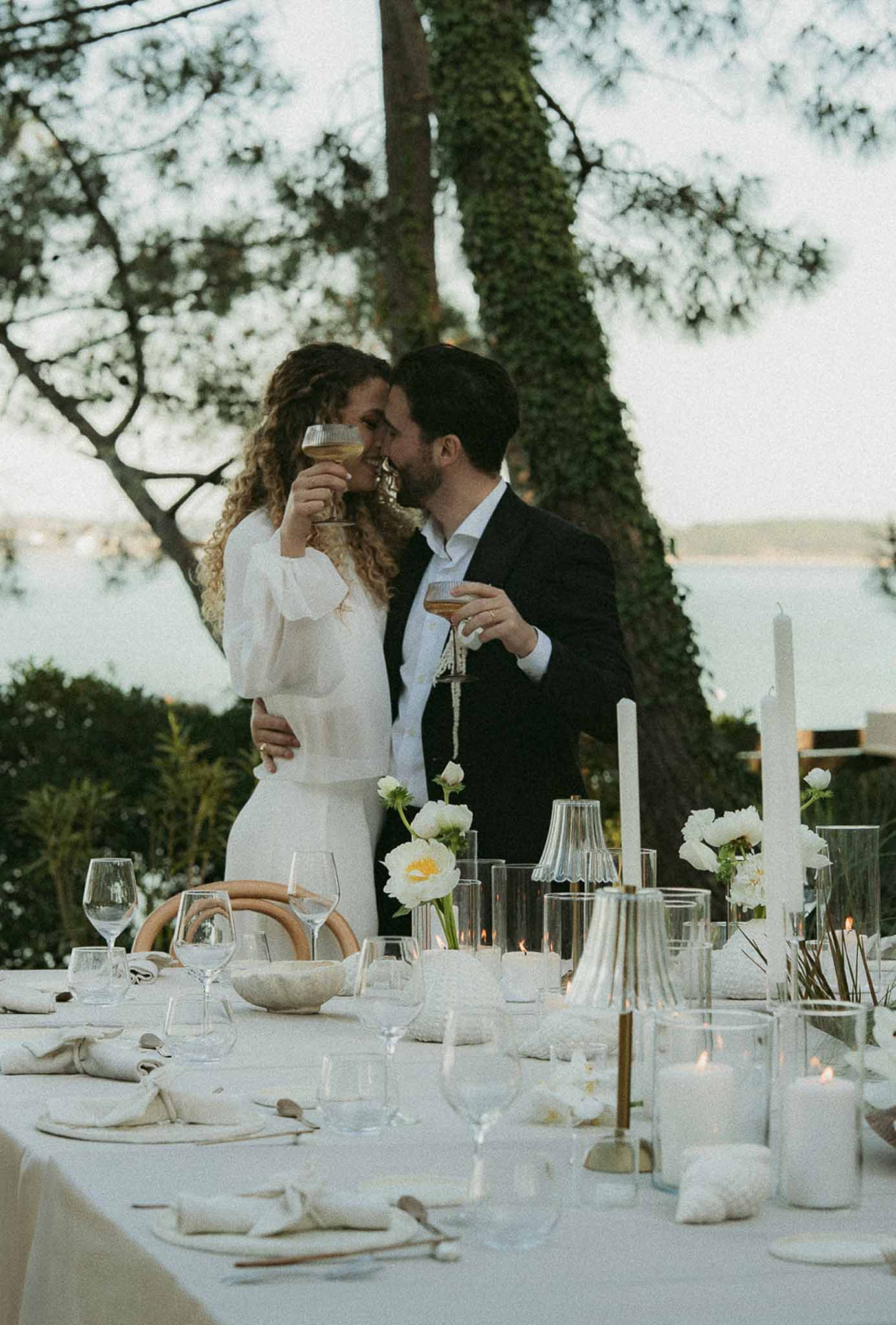 Couple toasting with champagne coupes at candlelit outdoor reception table with white peonies and rattan chargers