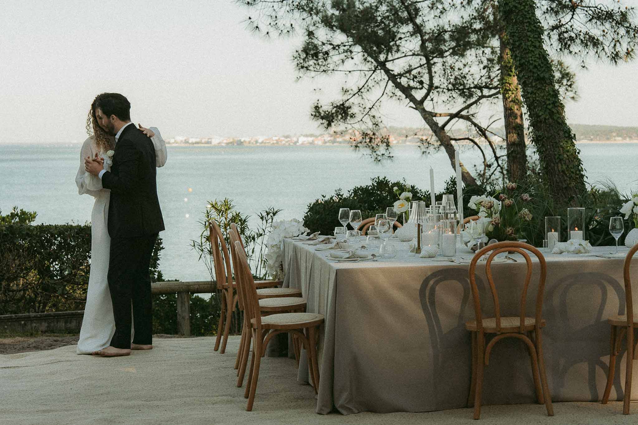 Couple sharing slow dance on seaside terrace at dusk beside candlelit reception table with white floral arrangements