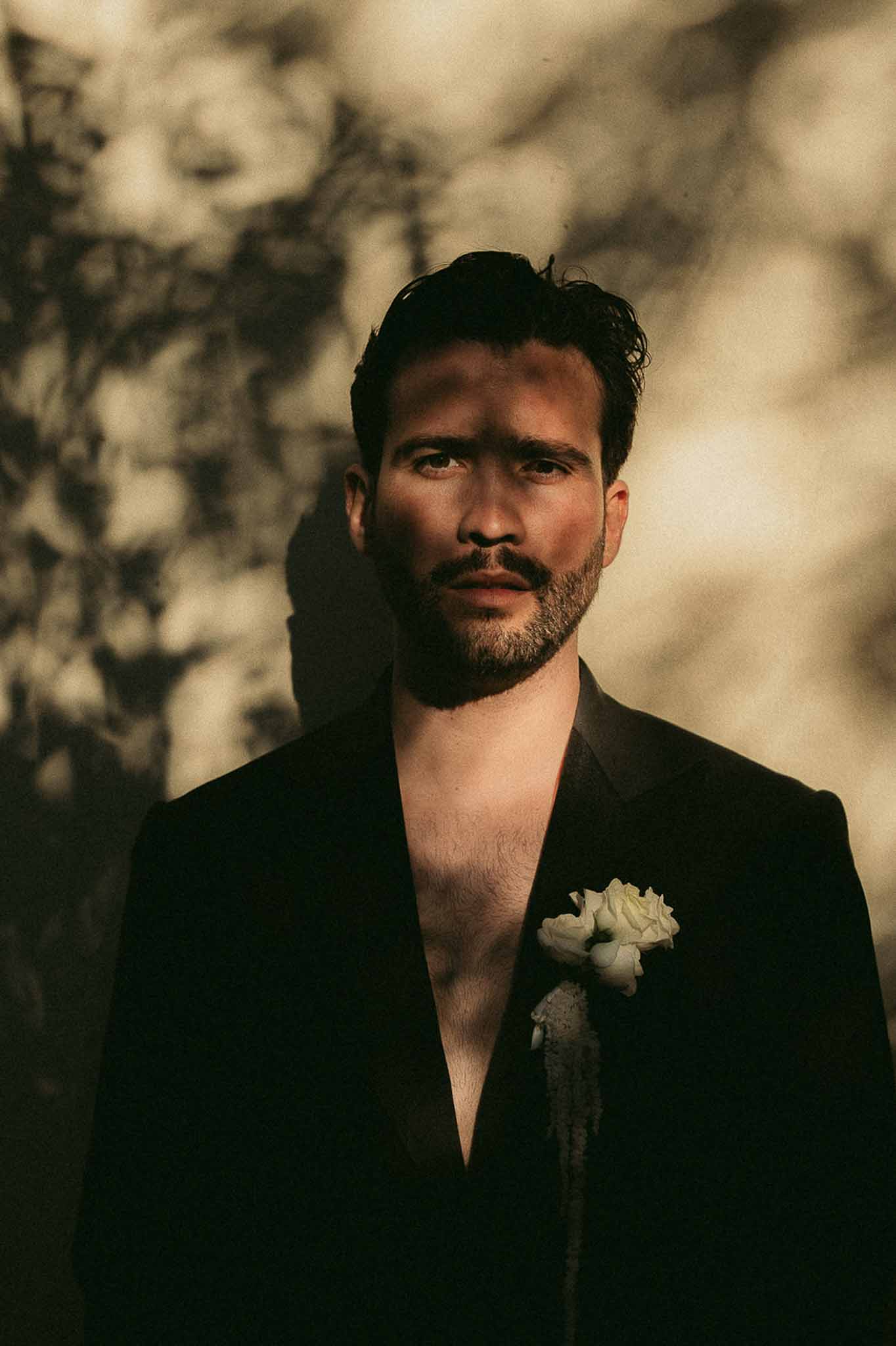 Groom in black suit with no shirt and white rose boutonniere in warm golden light with shadow patterns