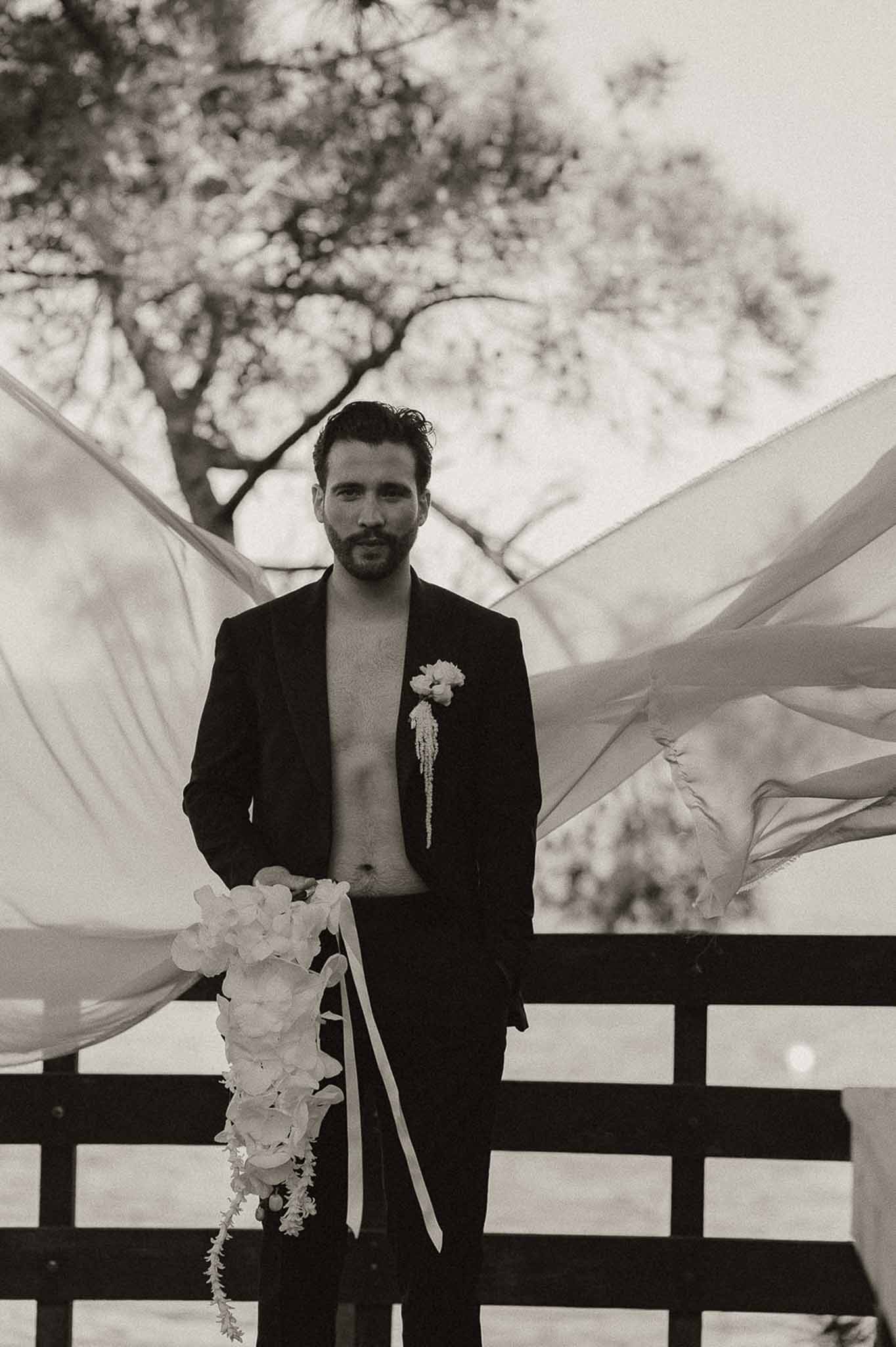 Black and white portrait of groom in open jacket holding cascading hydrangea bouquet with ribbon streamers