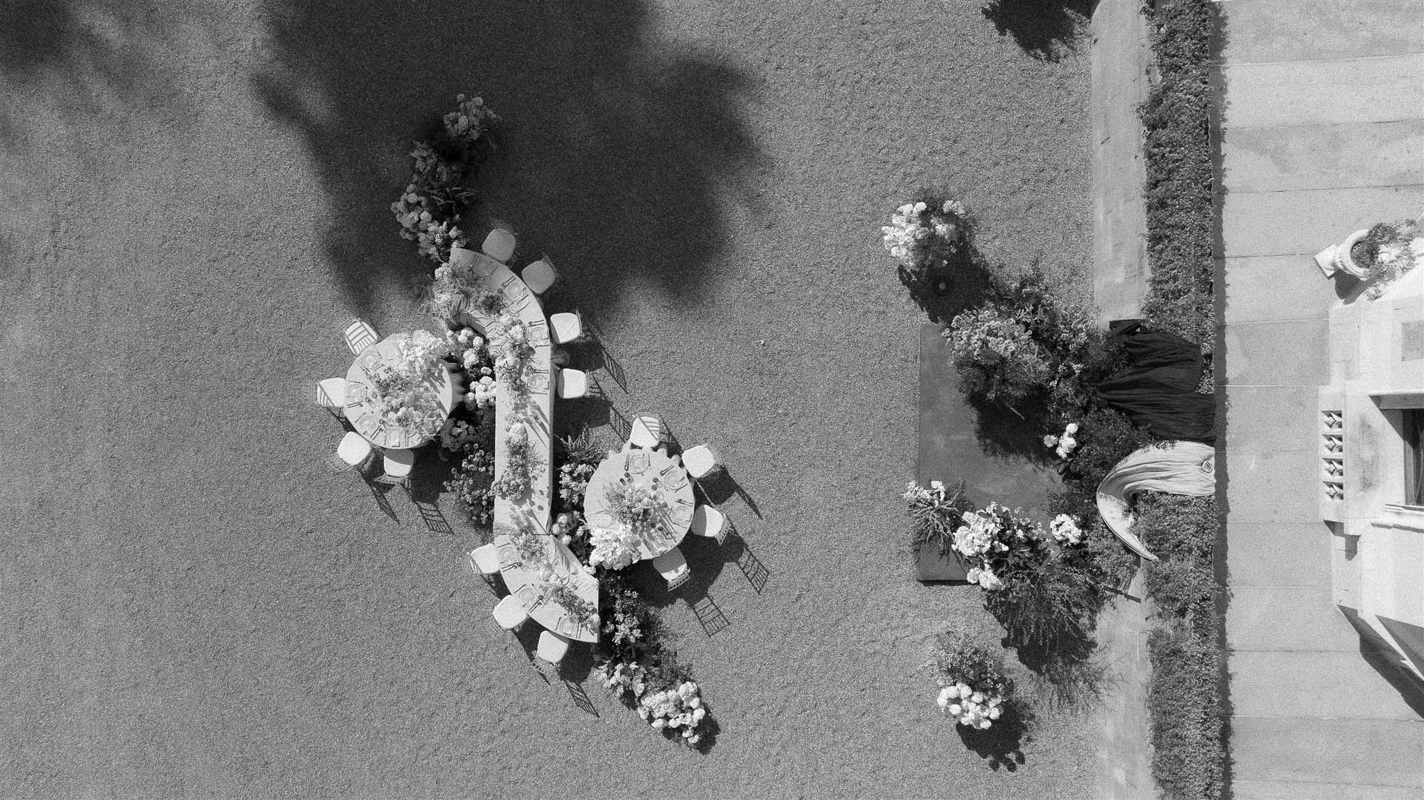Black-and-white aerial view of outdoor reception with three round tables, floral ground runners, and palm tree shadows on ...