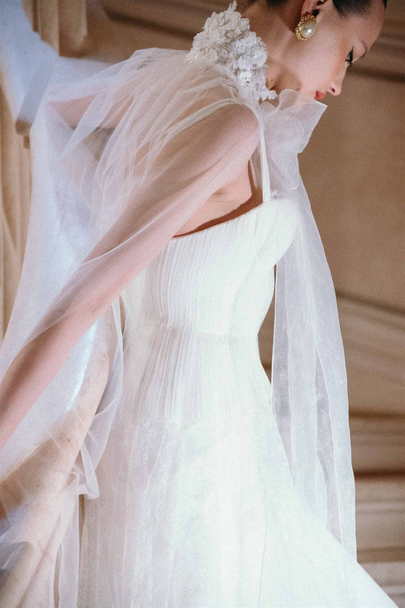 Bride in ivory pleated gown with sheer floral-applique cape and gold pearl drop earrings in chateau interior