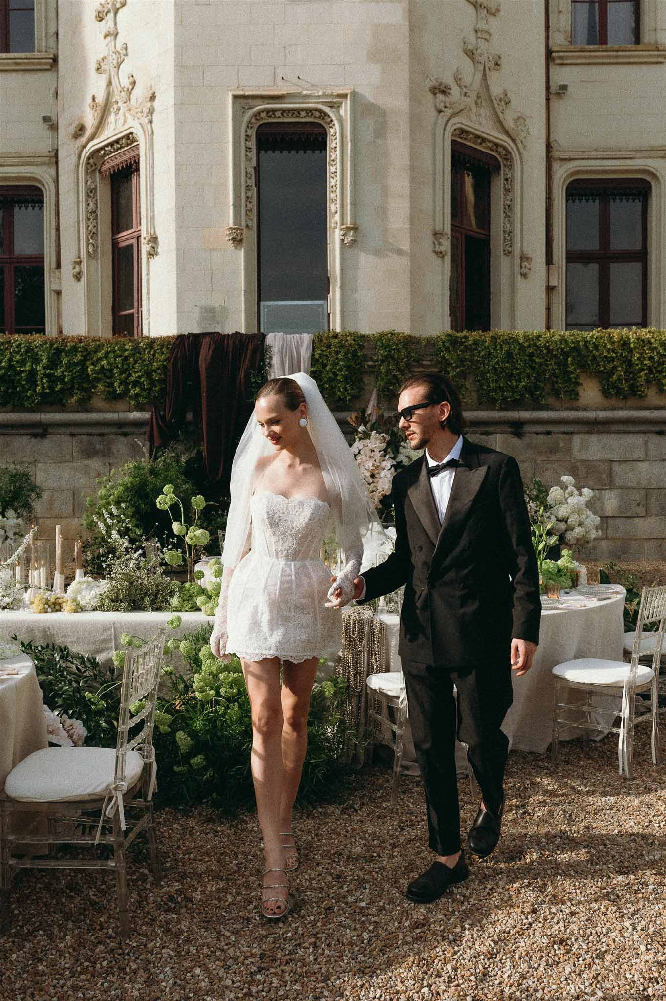 Bride in lace mini dress with veil and groom in tuxedo walk past green viburnum reception tables