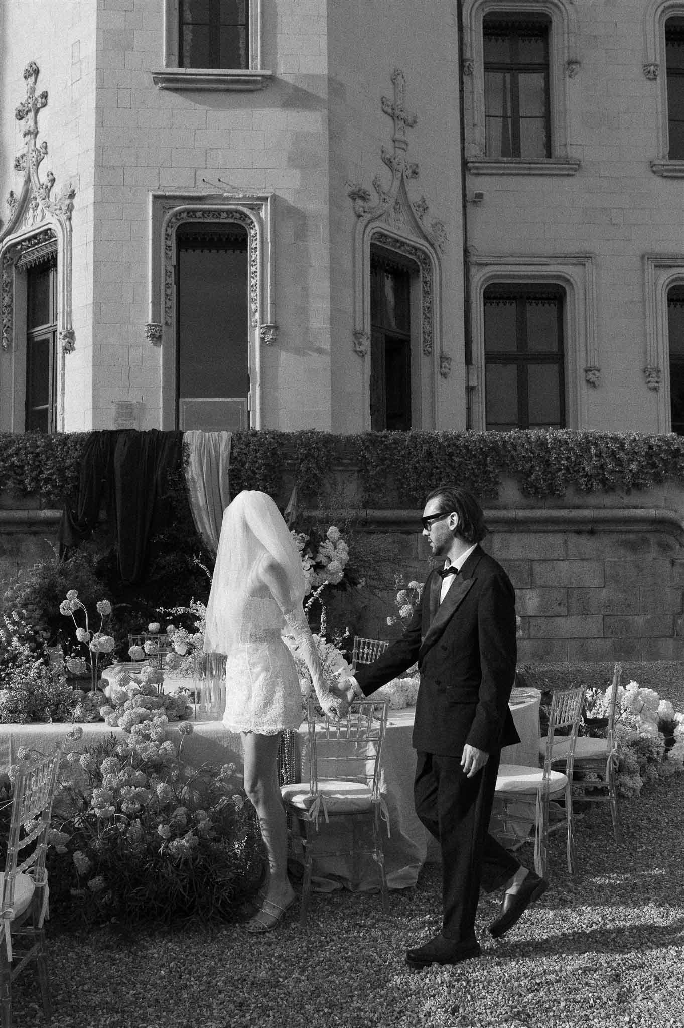 Black and white of couple in short lace dress and tuxedo walking past floral reception tables at Gothic-carved chateau