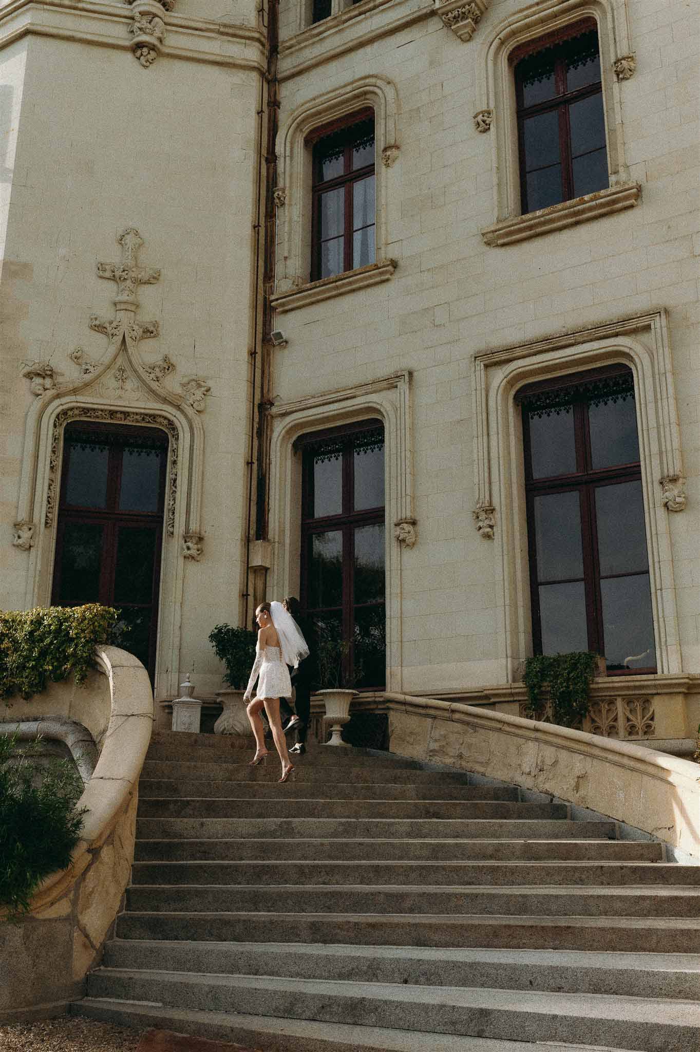 Bride in mini dress with flowing veil and groom walk up Gothic-Renaissance chateau stone staircase