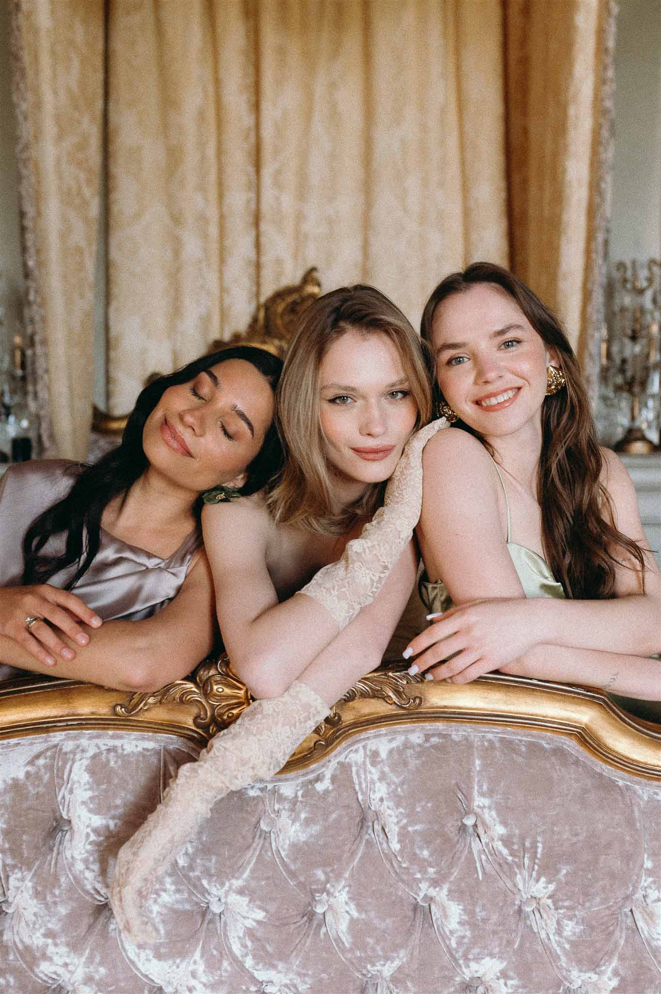 Bride with two bridesmaids in mauve and sage satin dresses leaning over blush velvet sofa in gilt-paneled chateau room