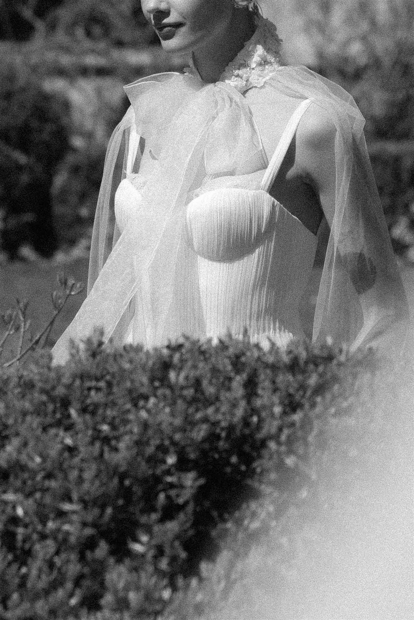 Black and white detail shot of bride wearing pleated bodice gown with sheer tulle cape draped over shoulders