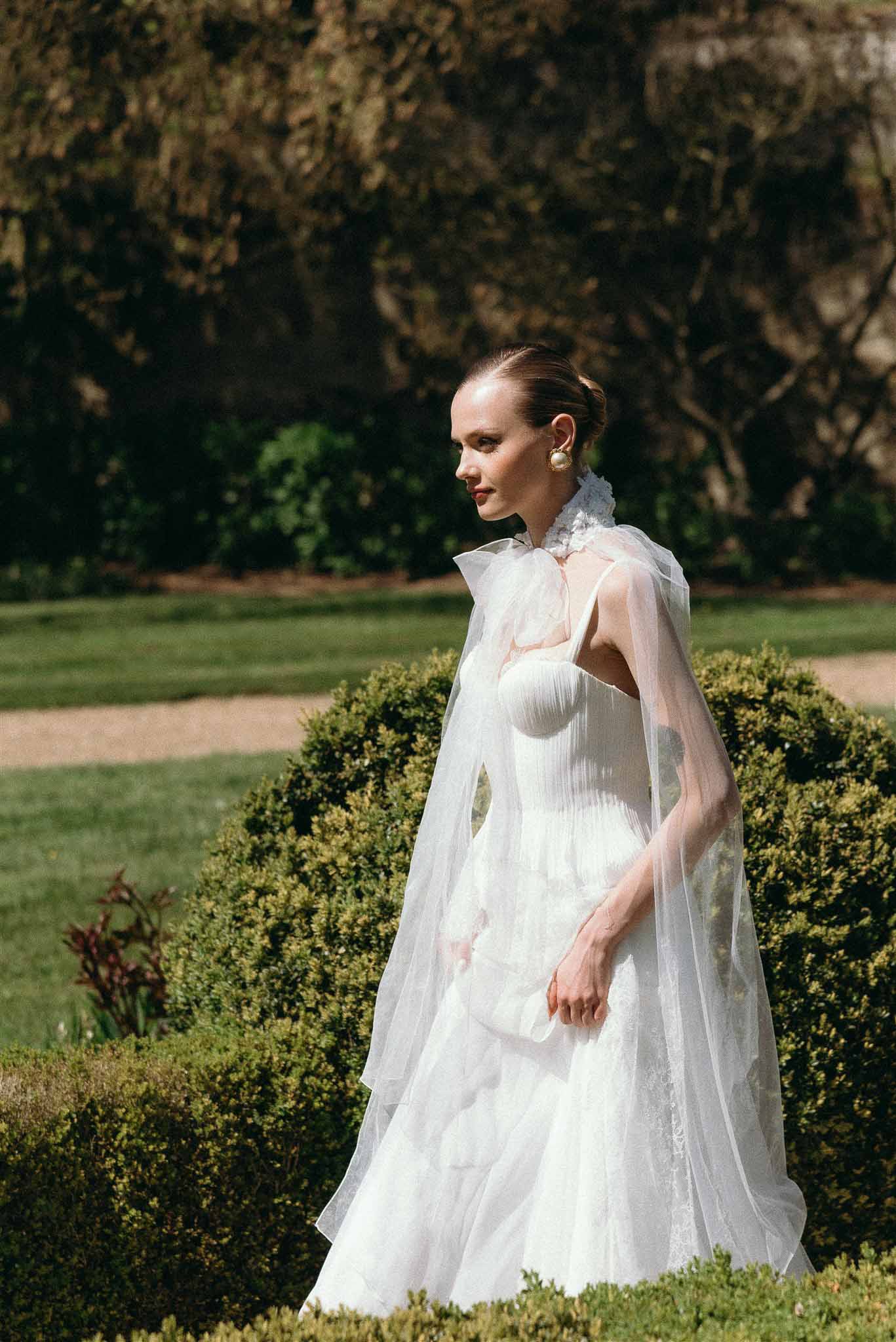 Bride in strapless white gown with sheer tulle cape posing among boxwood hedges in formal garden