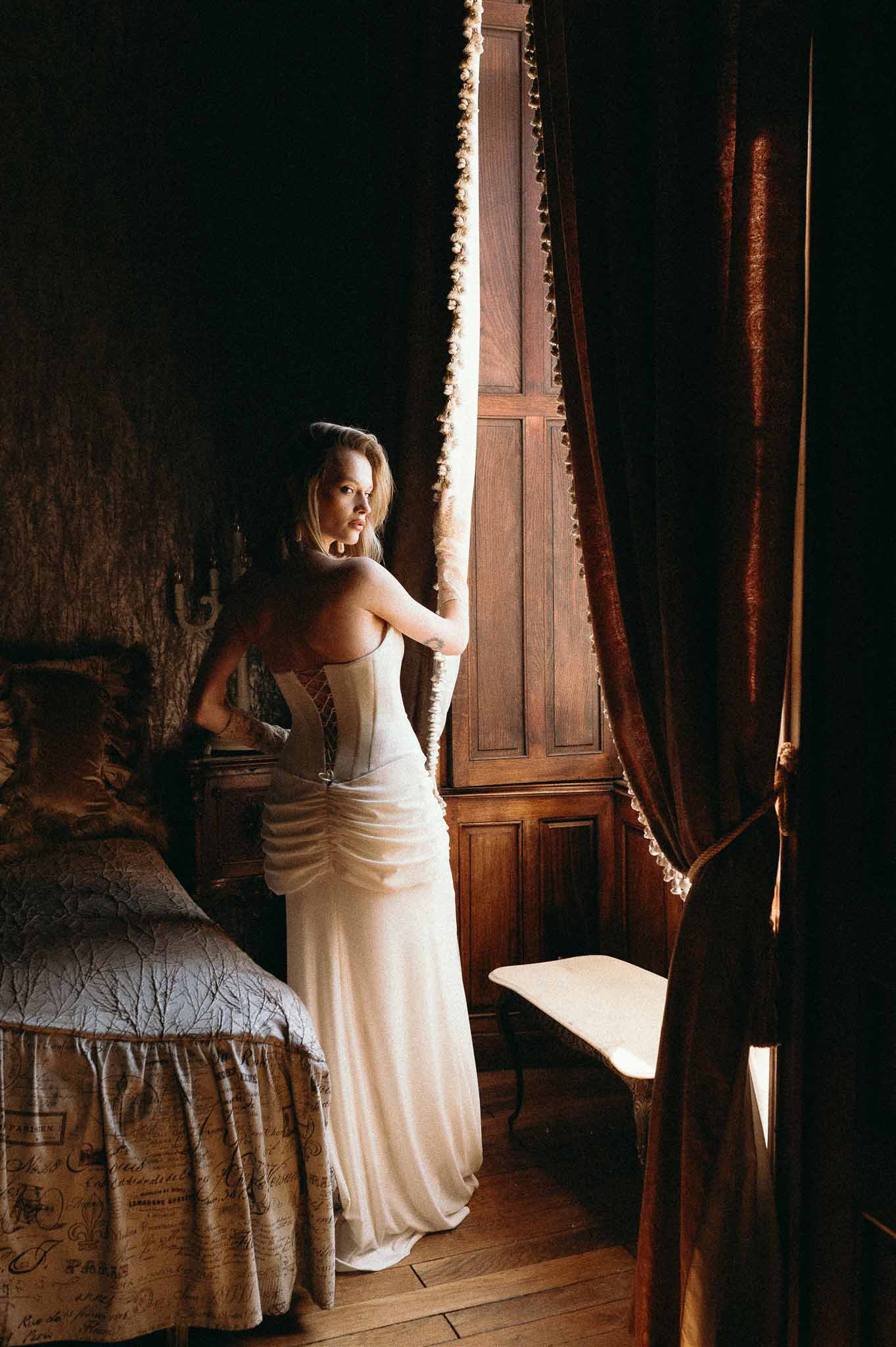 Bride in ivory ruched gown with corseted back looking over shoulder in dark chateau bedroom with velvet curtains