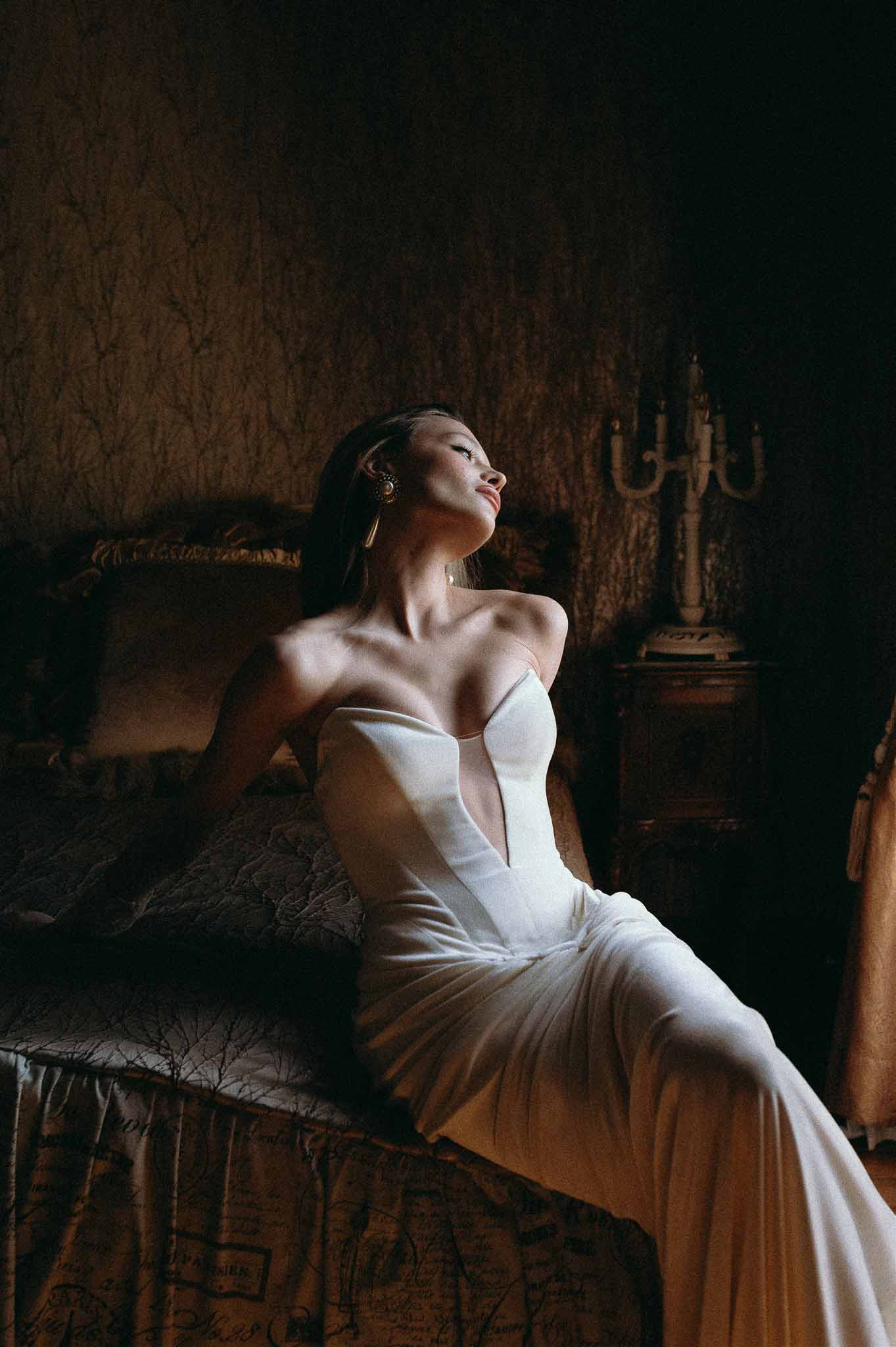 Bride in strapless ivory gown seated on antique bed with dramatic side lighting in botanical wallpaper room