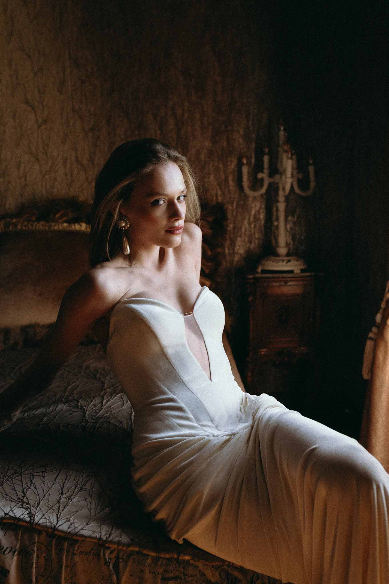 Bride in ivory V-neck satin gown with gold earrings seated on moody chateau bed with candelabra