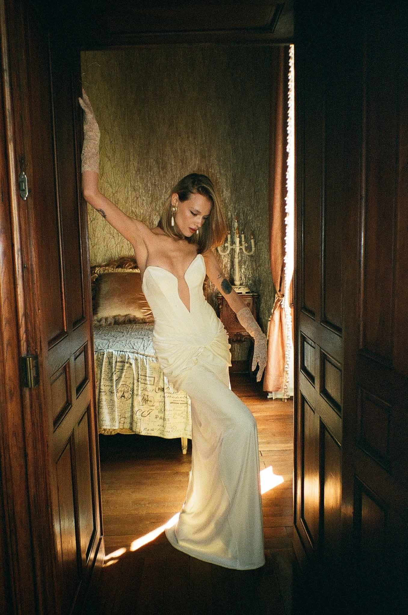 Bride in deep V-neck satin gown with lace gloves posing in doorway of vintage chateau bedroom