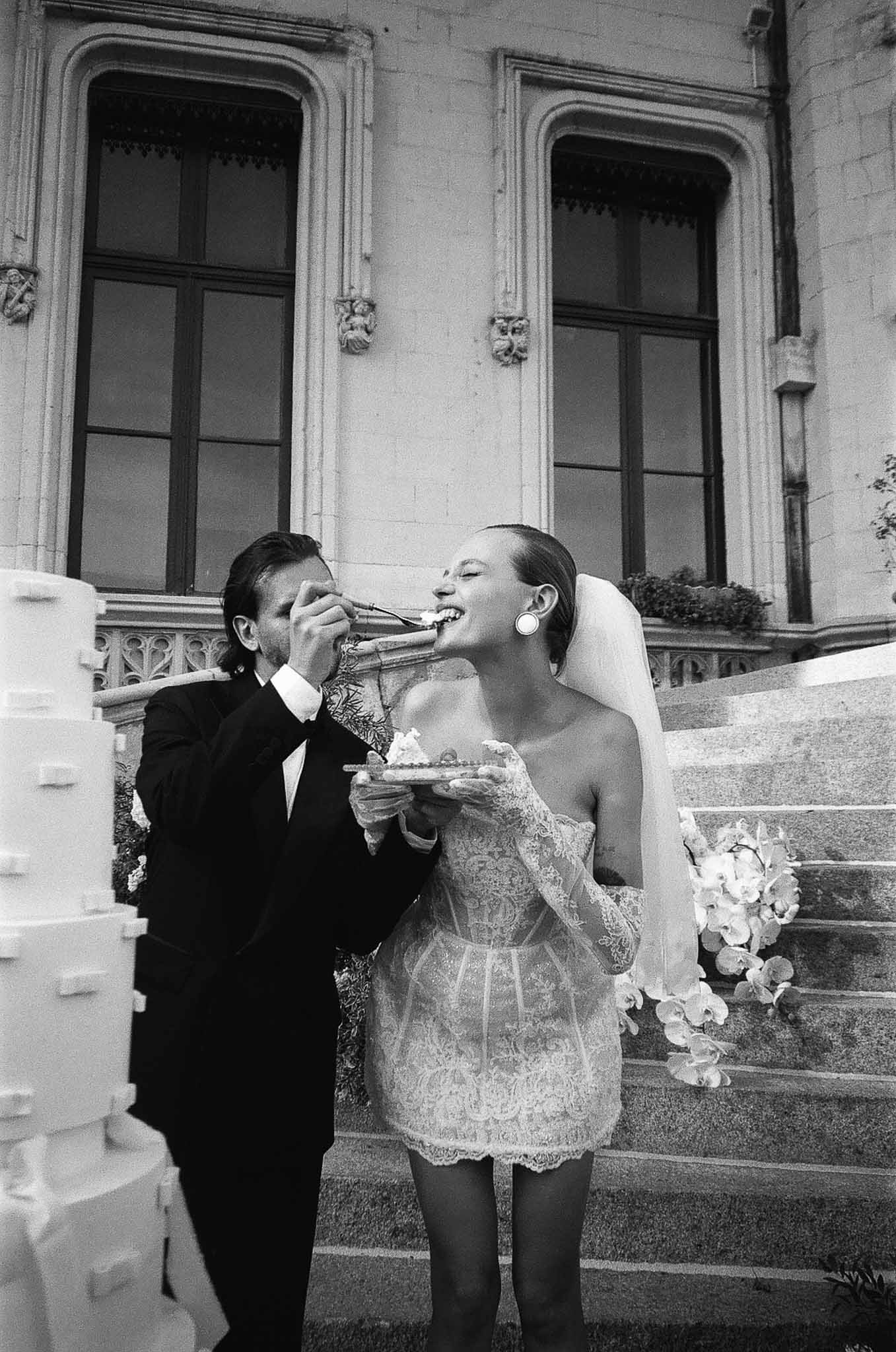 Black and white of groom feeding bride cake on chateau steps, bride in lace mini dress with orchid bouquet