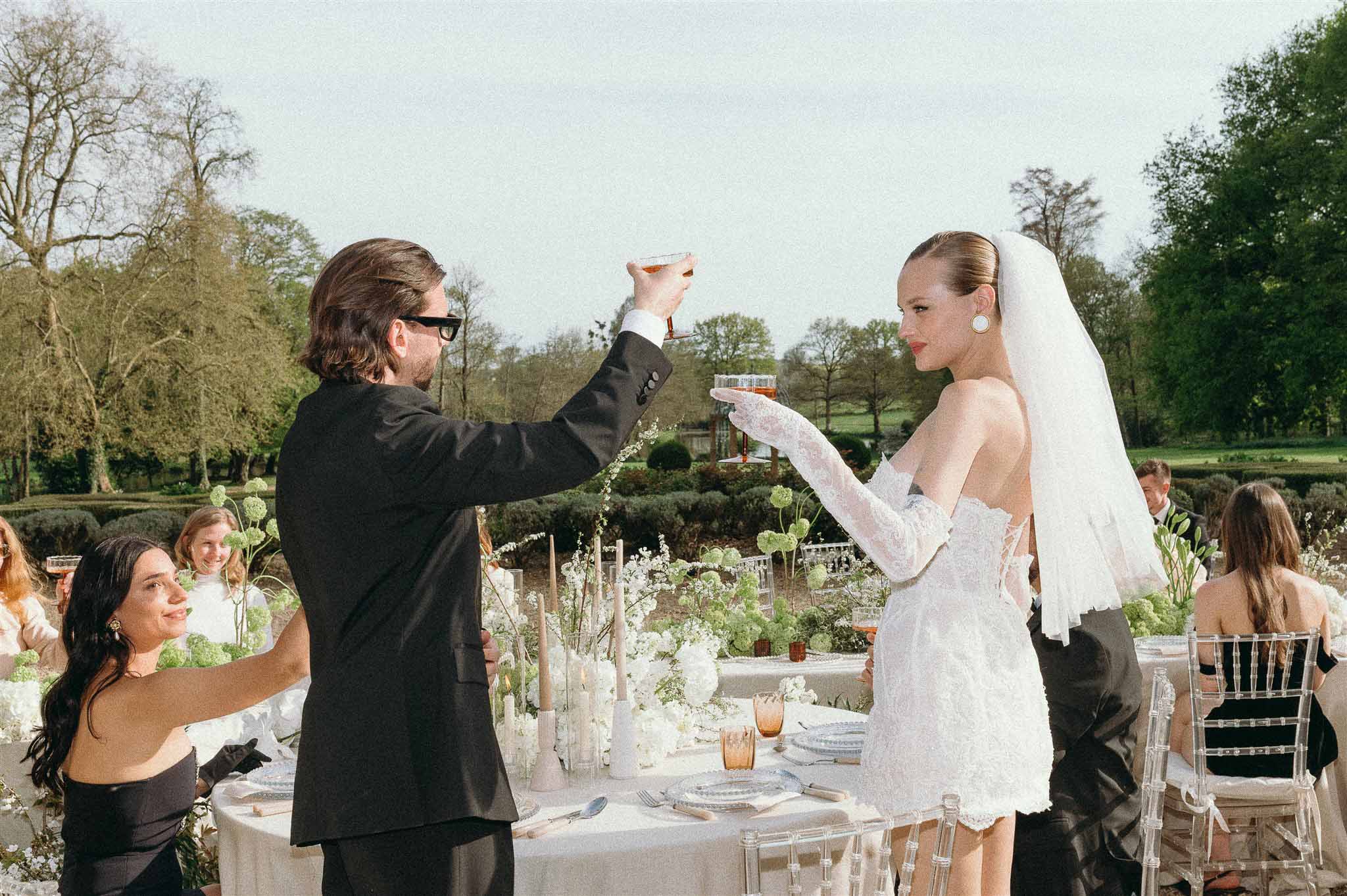 Couple toasting at round table with viburnum and white blooms, ghost chairs, and topiary garden behind