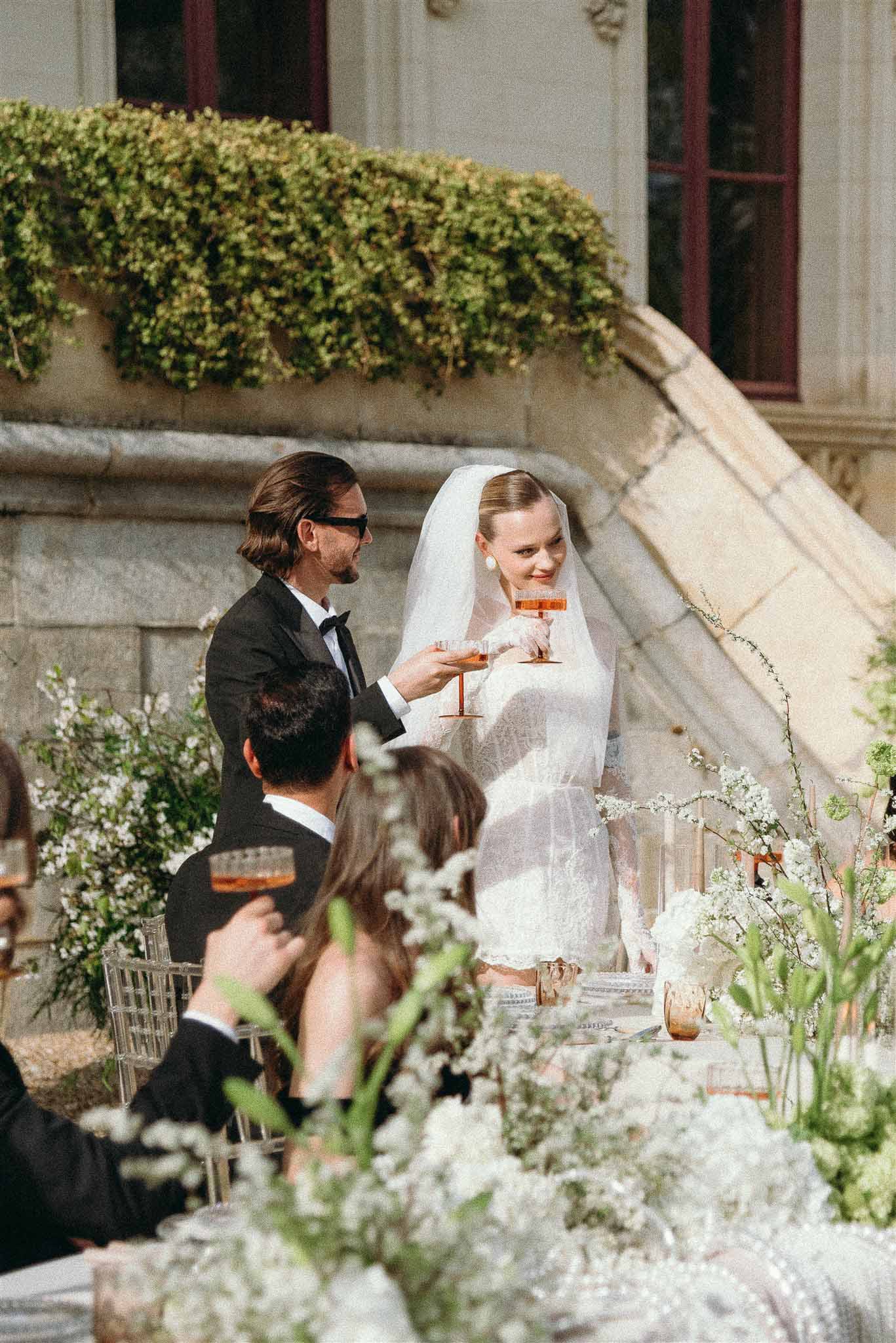 Bride in white lace dress and groom in tuxedo clinking coupe glasses at outdoor chateau reception table