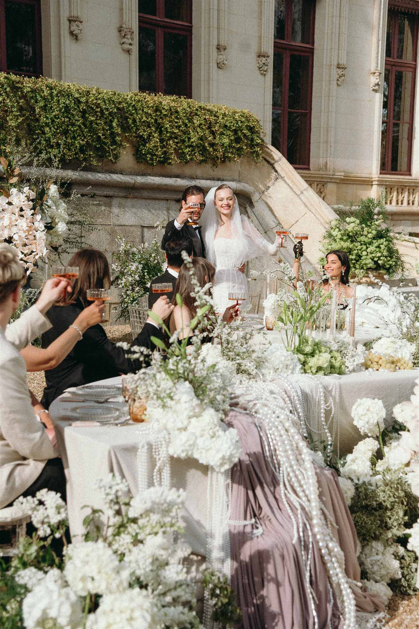 Couple toasting with guests at white hydrangea and blossom tables with pearl garlands before carved stone chateau