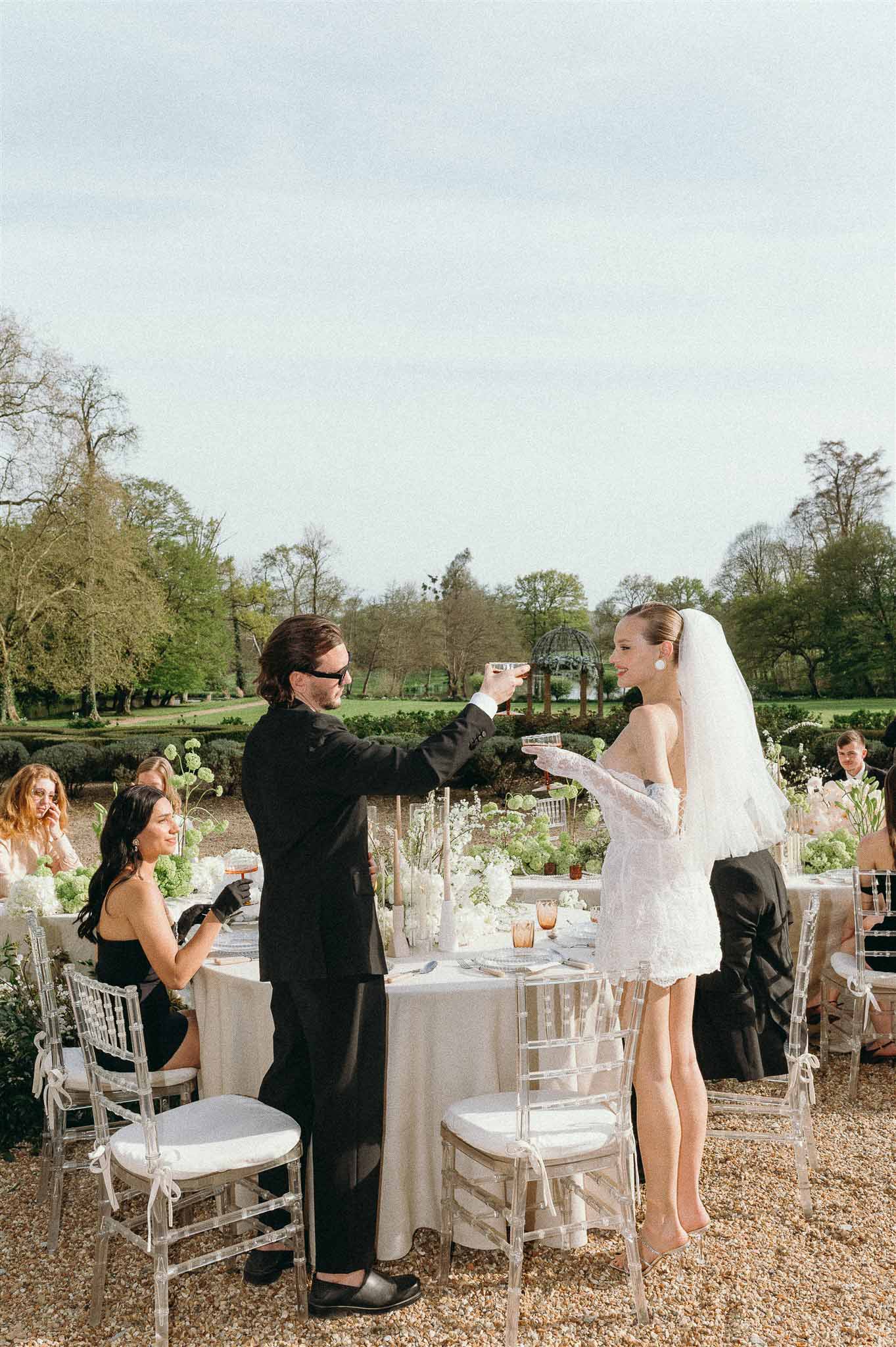Couple in lace mini dress and black suit toasting at ghost-chair table with green viburnum and white tulips