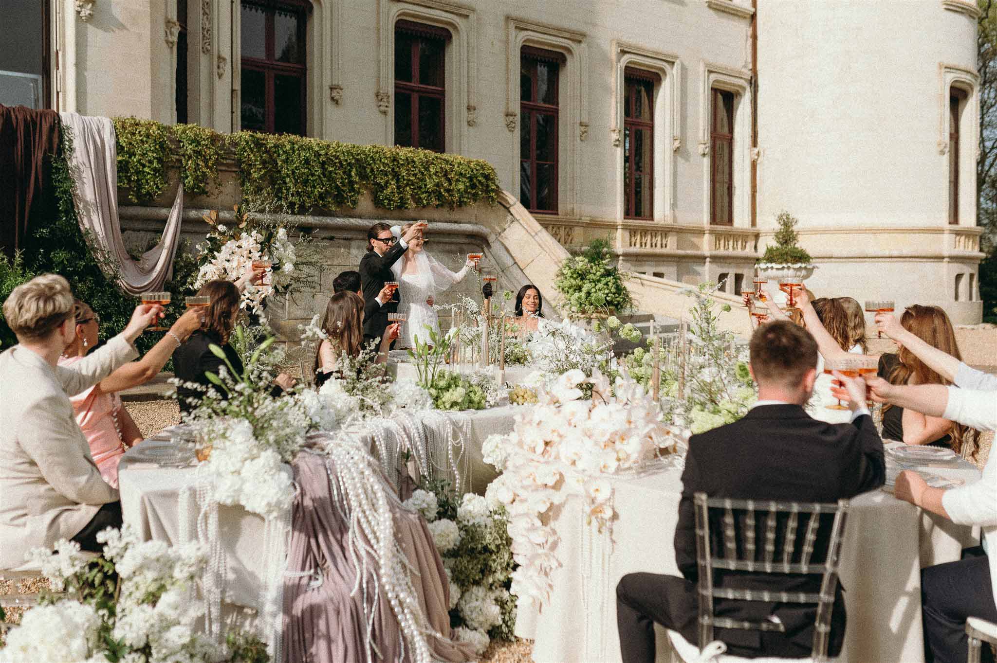 Bride and groom toast with guests at orchid and hydrangea table before chateau facade in afternoon sun