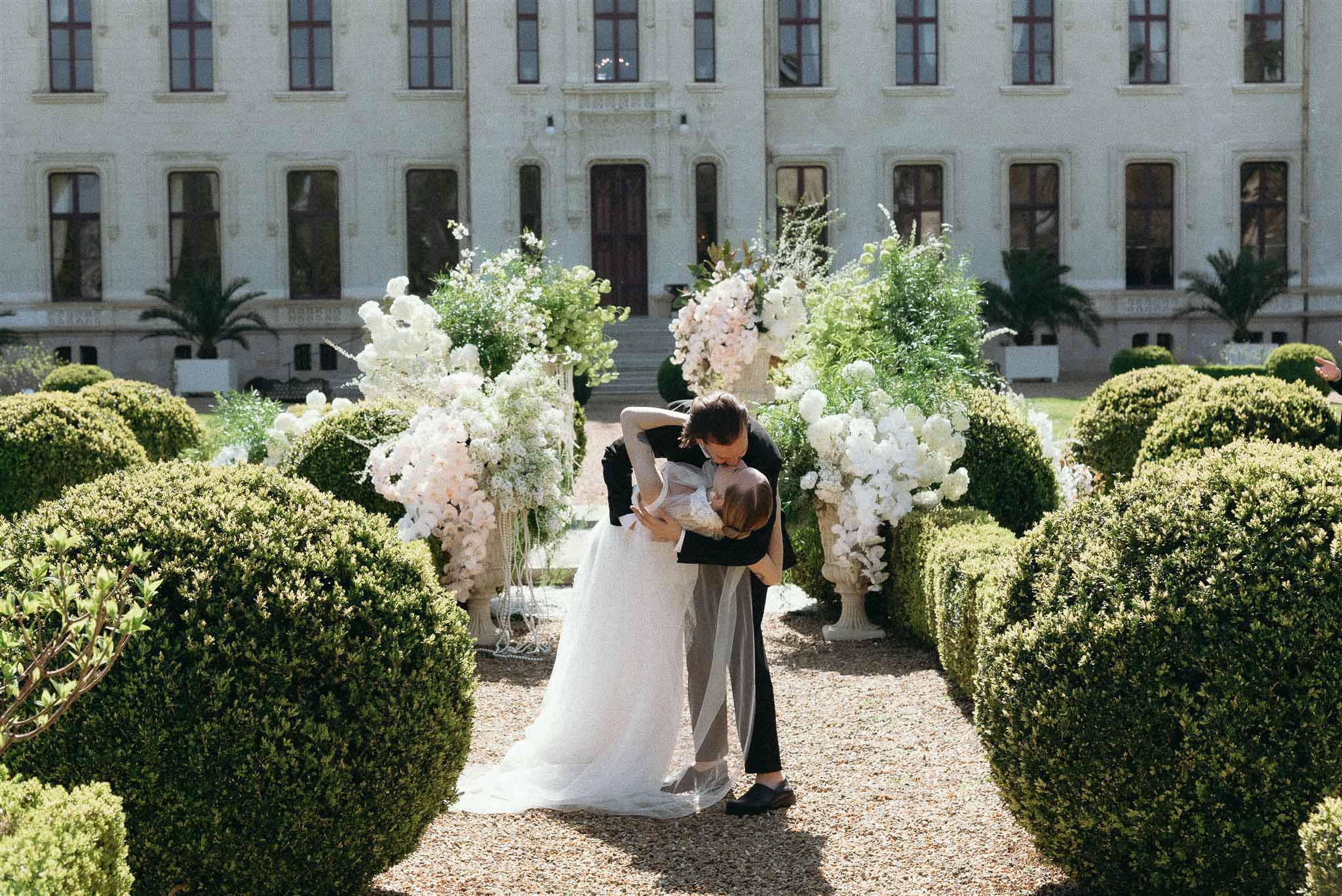 Couple dip kiss on garden path flanked by white orchid urns and rounded topiary before chateau