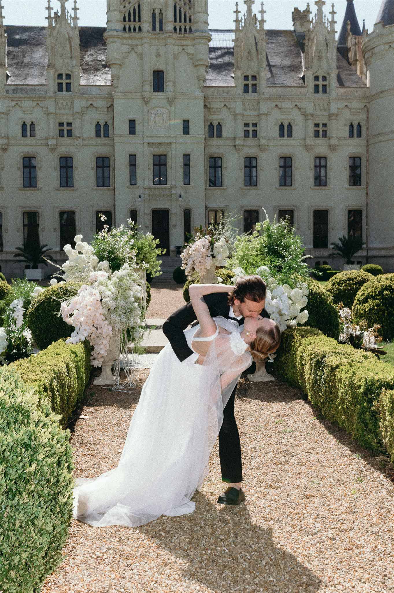 Groom dipping bride for kiss on garden path with white floral urns and Gothic chateau behind