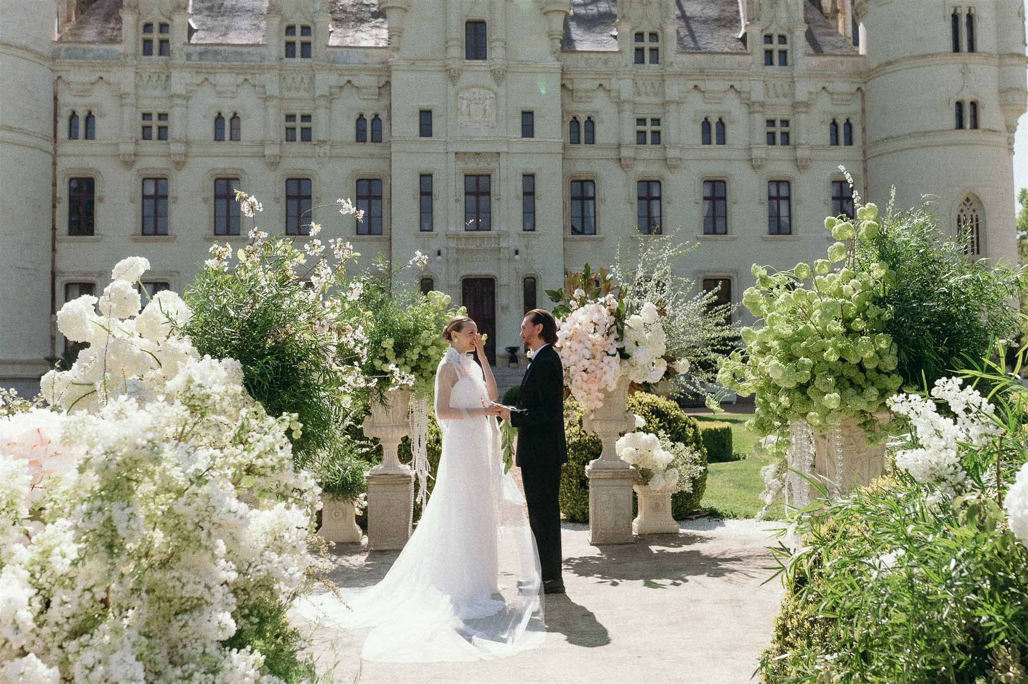 Bride and groom exchange vows at outdoor chateau ceremony flanked by white hydrangea and peony urns