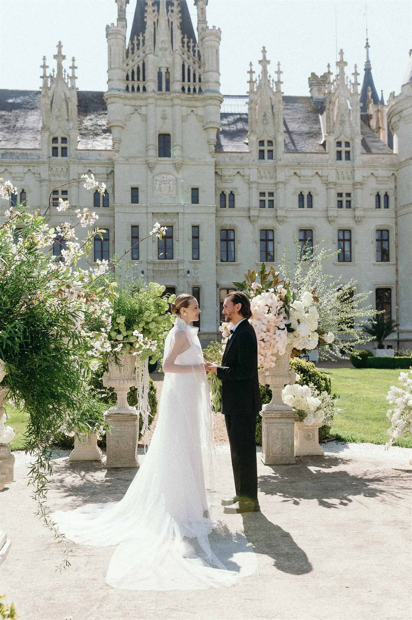 Outdoor ceremony at Gothic chateau with couple exchanging vows between white and blush floral urns