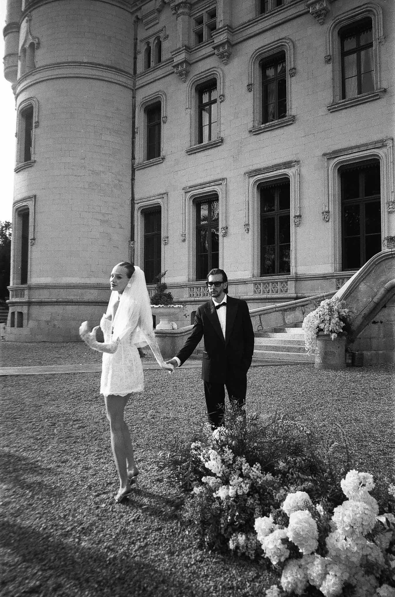 Black and white photo of couple walking across chateau courtyard with Renaissance facade and ground-level floral display