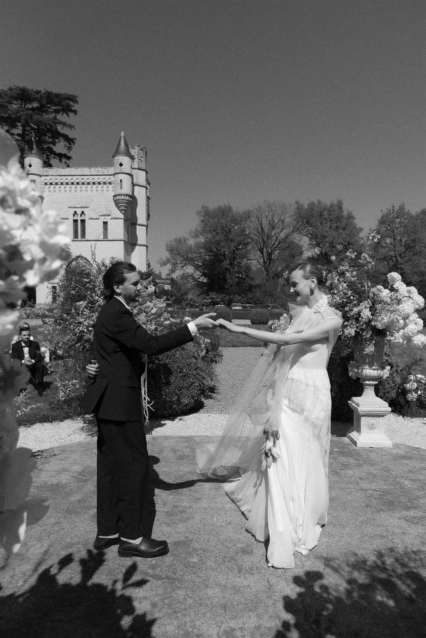 Couple twirls on gravel beside hydrangea urn with Gothic-tower chateau and sculpted hedges in B&W