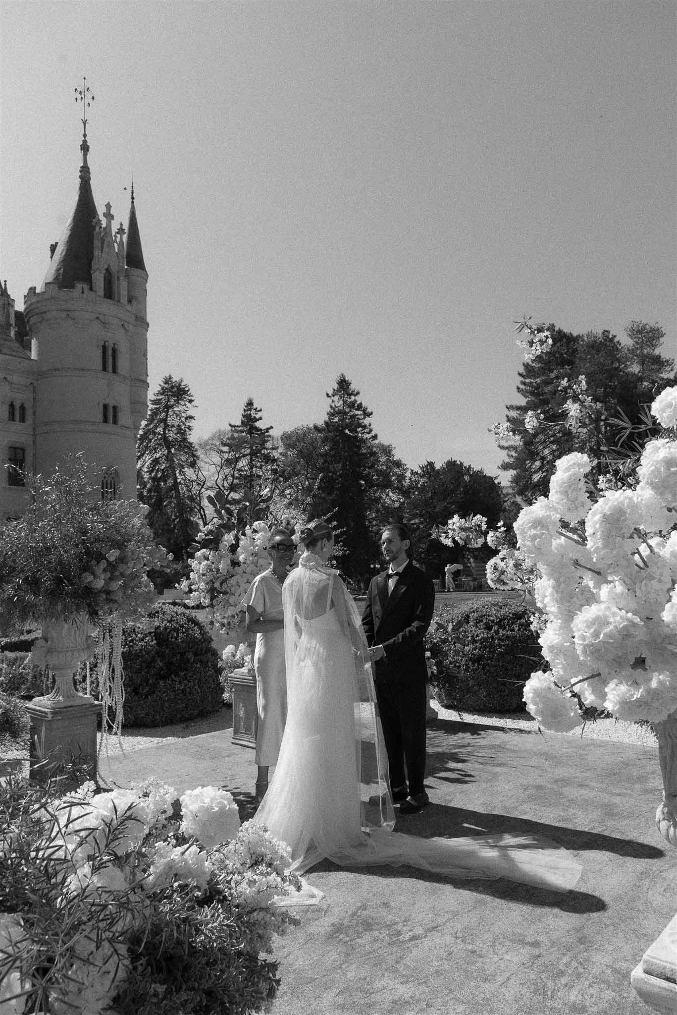 Black-and-white outdoor ceremony with bride and groom before officiant flanked by large floral arrangements at Gothic chateau