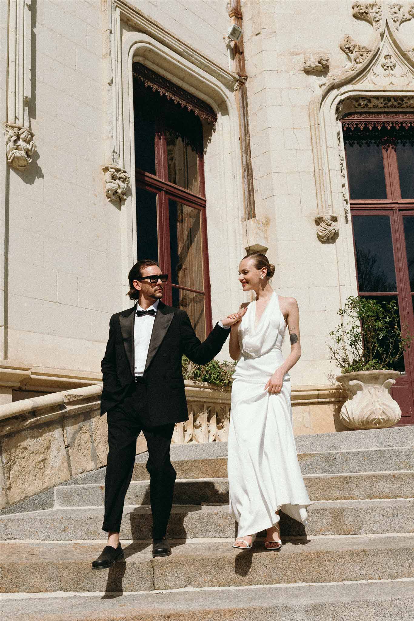 Bride in ivory halter-neck satin gown and groom in black tuxedo holding hands on carved stone chateau steps