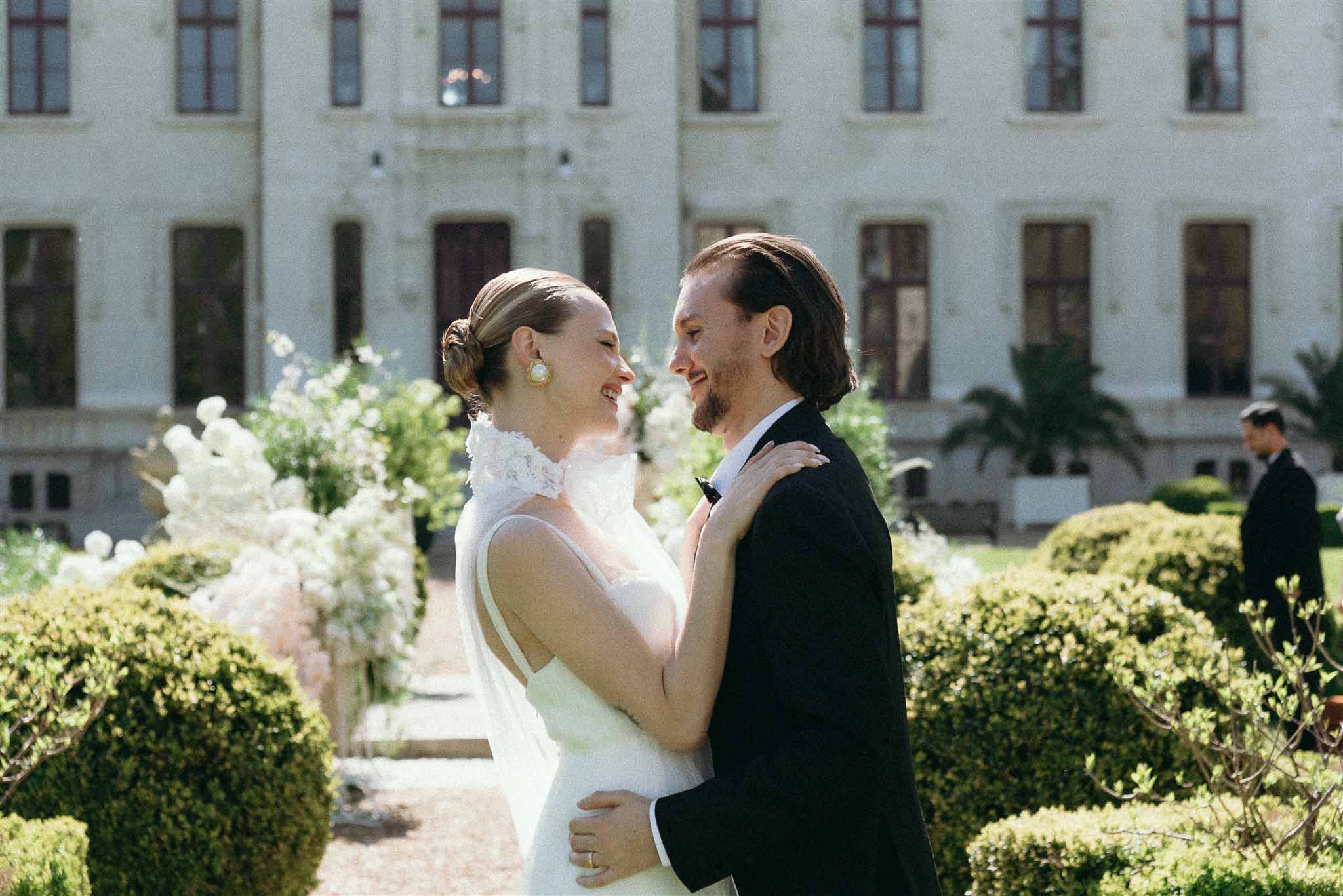 Bride in ivory gown with lace collar and groom in black suit embracing in formal chateau garden