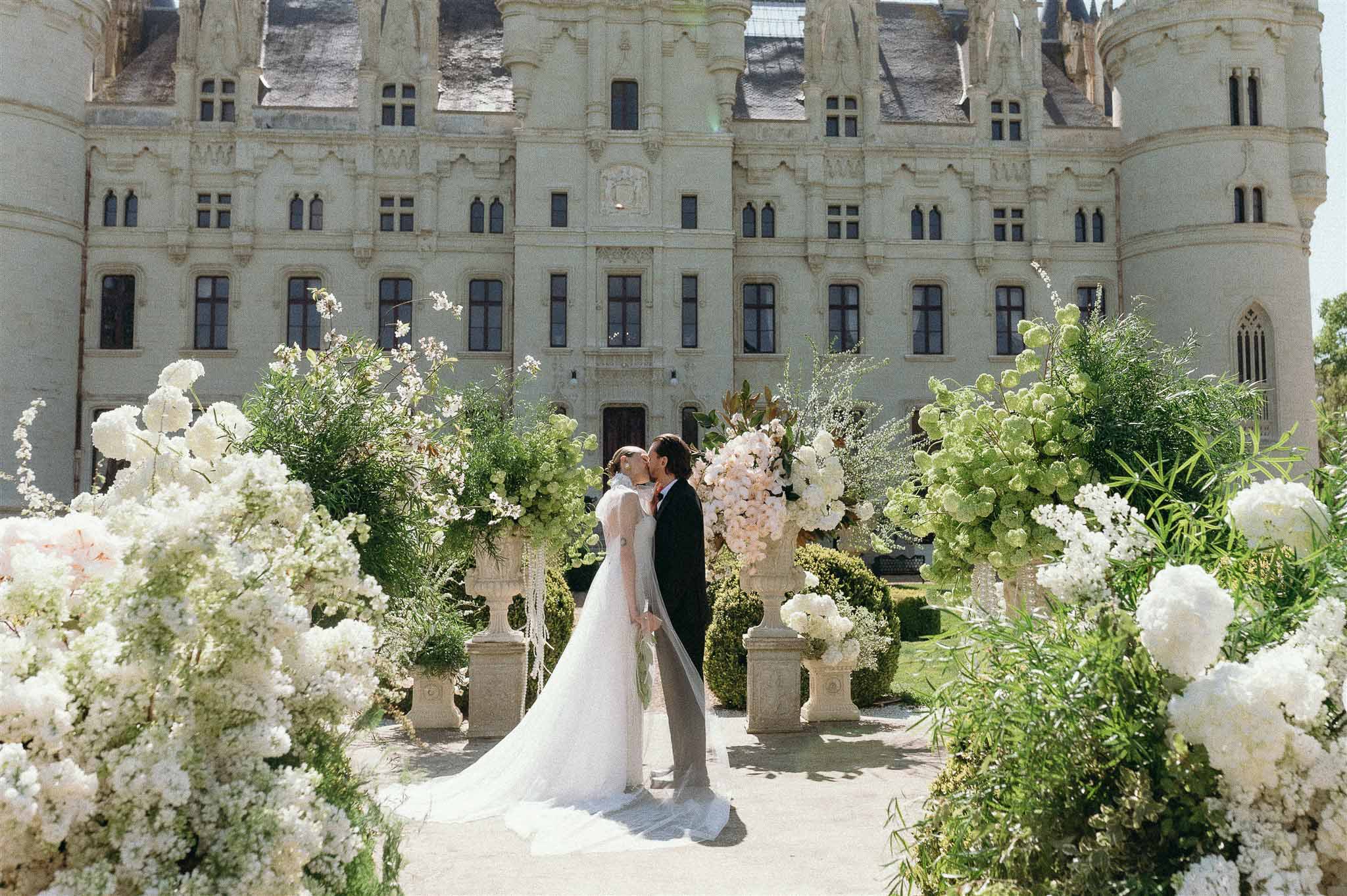 Couple kissing through white hydrangea and orchid urn aisle before neo-Gothic chateau with turrets and arched windows