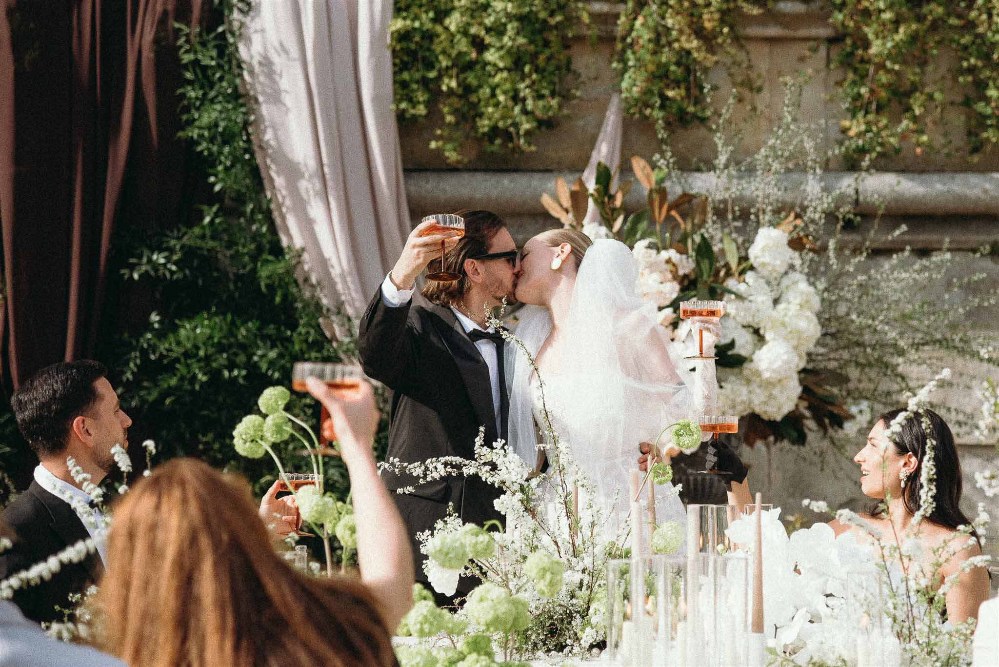 Bride and groom kissing while raising coupe glasses at reception with white hydrangea and greenery backdrop