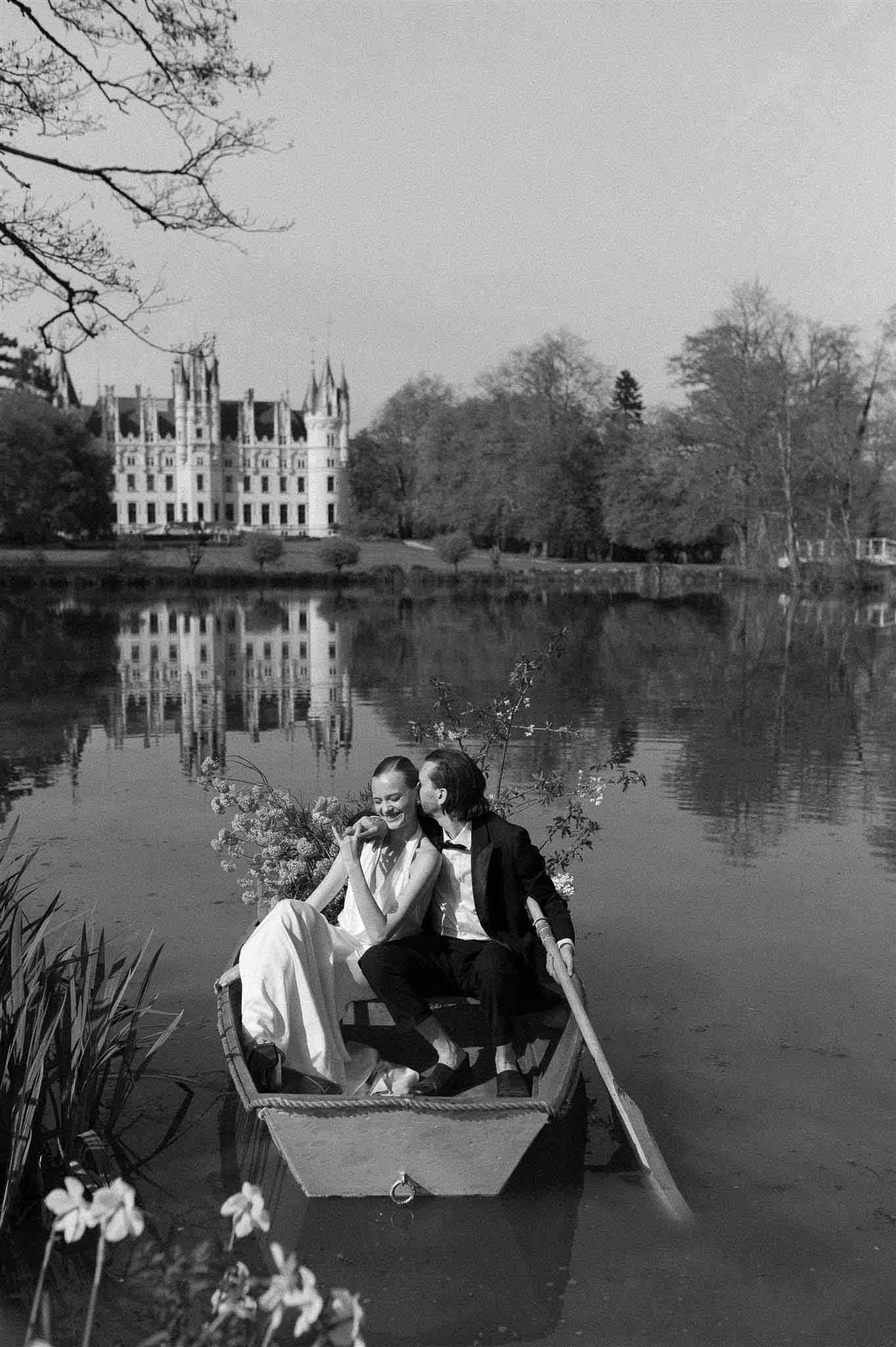 Black and white photo of bride and groom in rowboat on lake with French Renaissance chateau reflected in water