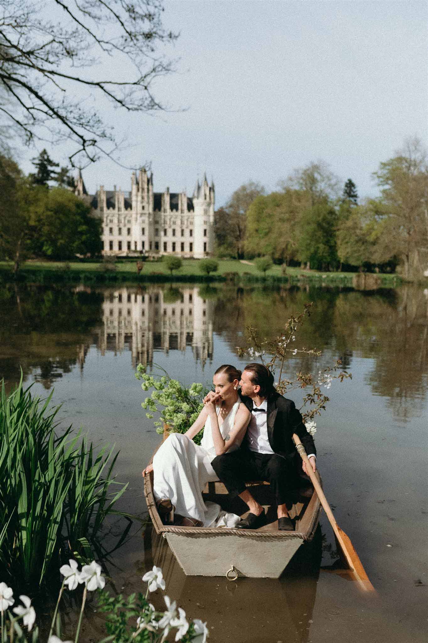 Couple in wooden rowboat on lake before white Gothic chateau with narcissus flowers and green foliage