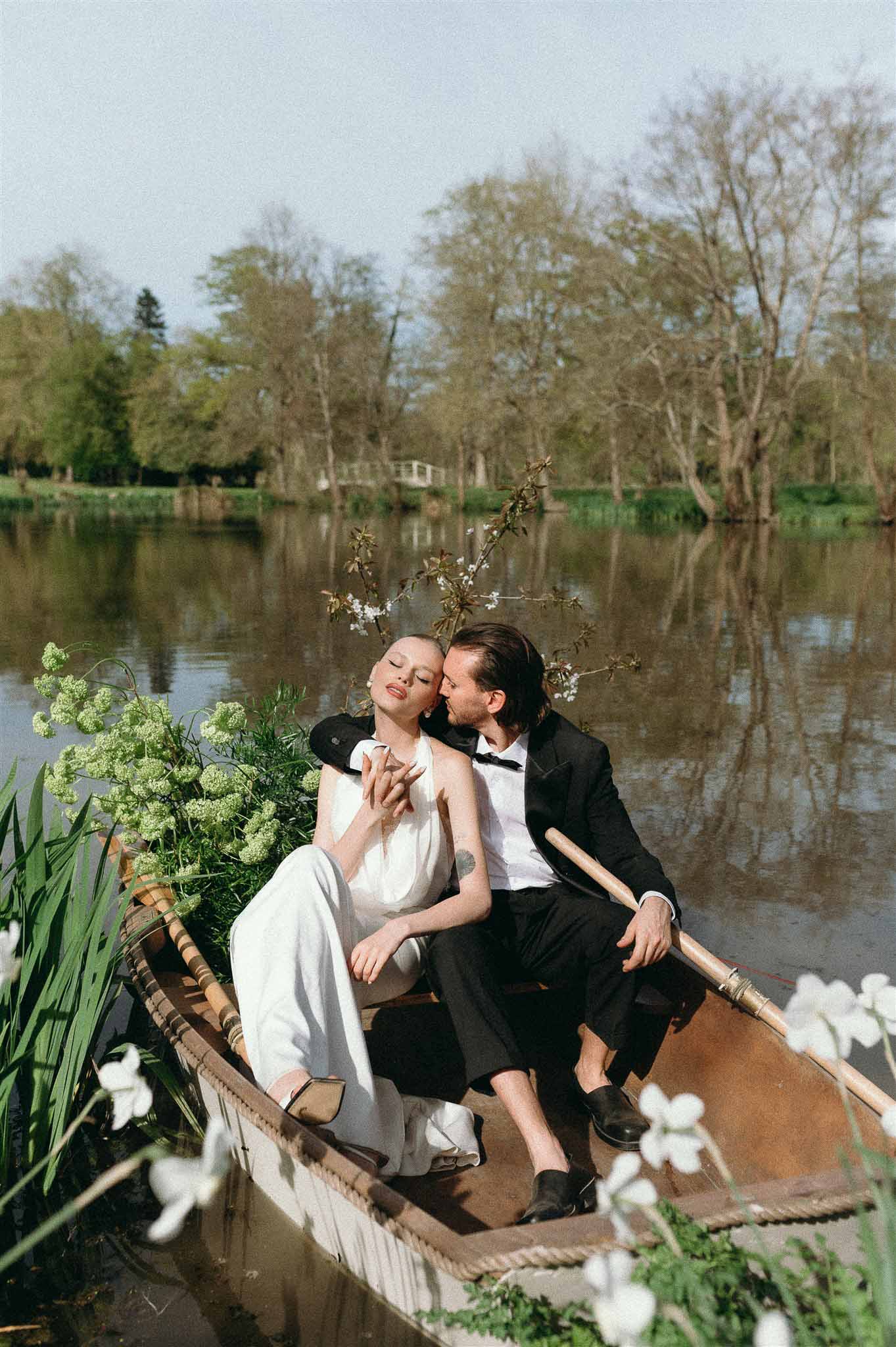 Bride in white bridal jumpsuit and groom in black tuxedo seated in flower-filled rowboat on calm river