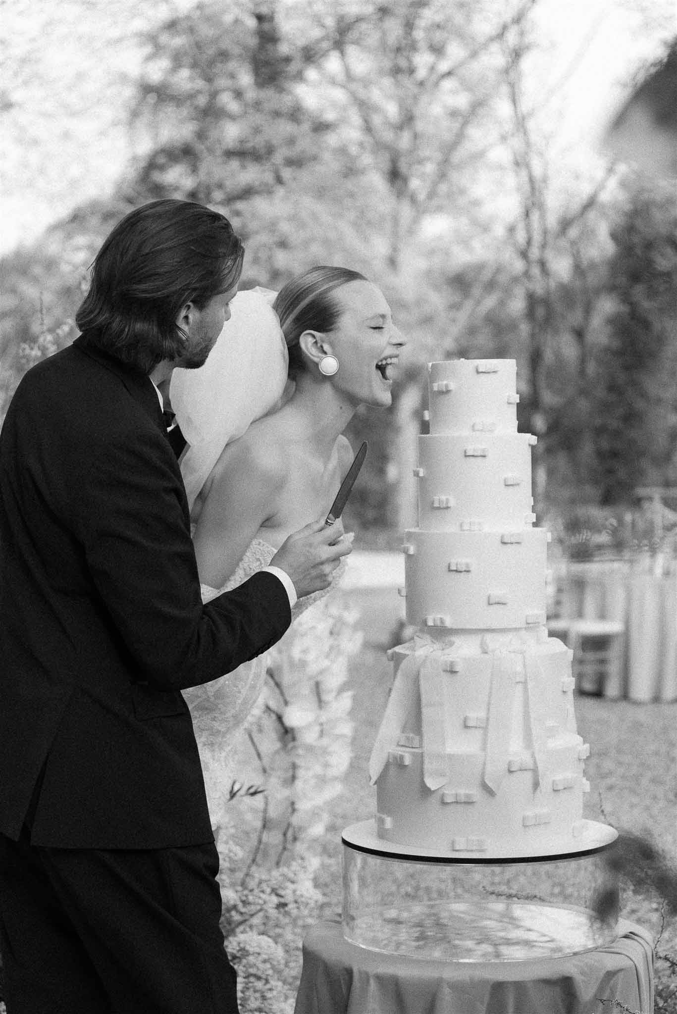 Bride and groom cutting four-tier wedding cake decorated with bow motifs outdoors, black and white photo