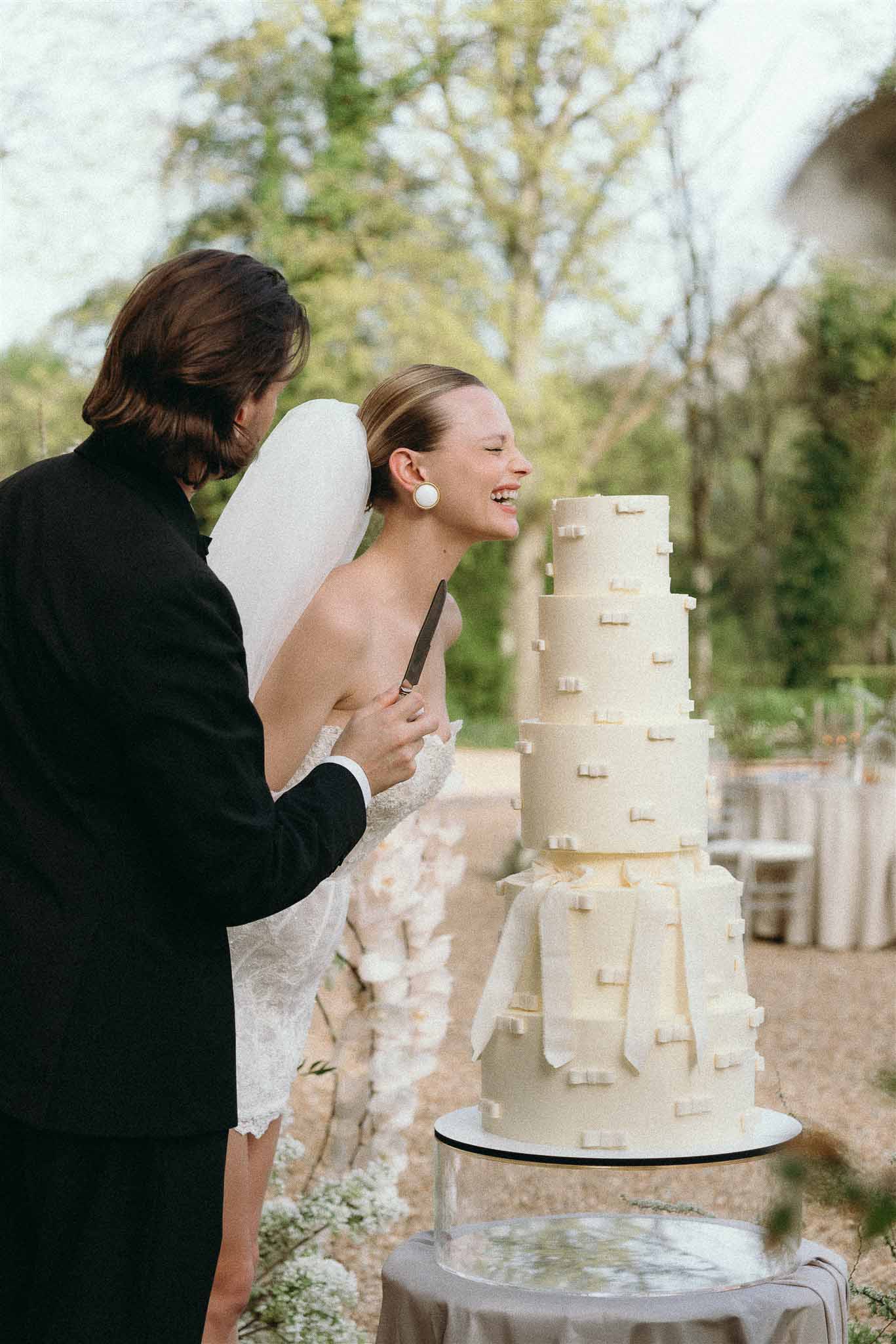 Bride laughing as couple cuts four-tier ivory cake with fondant bow decorations on mirrored stand at outdoor reception